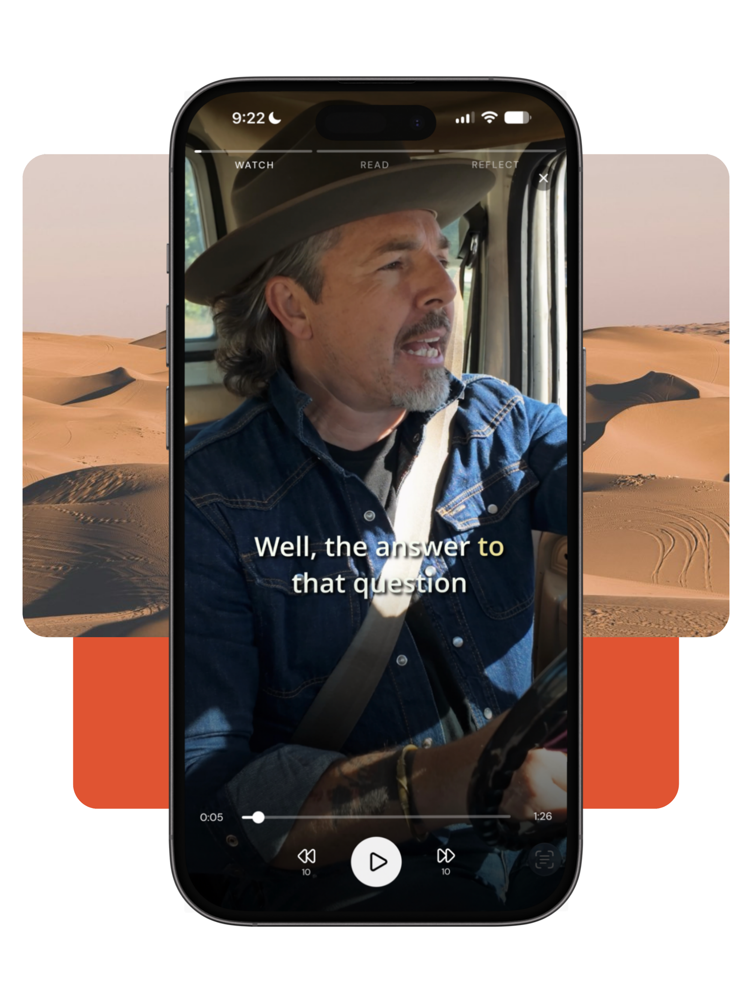 an iPhone showing a man with gray hair, a beard, wearing a cowboy hat and denim jacket, smiling while driving a vehicle in a desert landscape with sand dunes