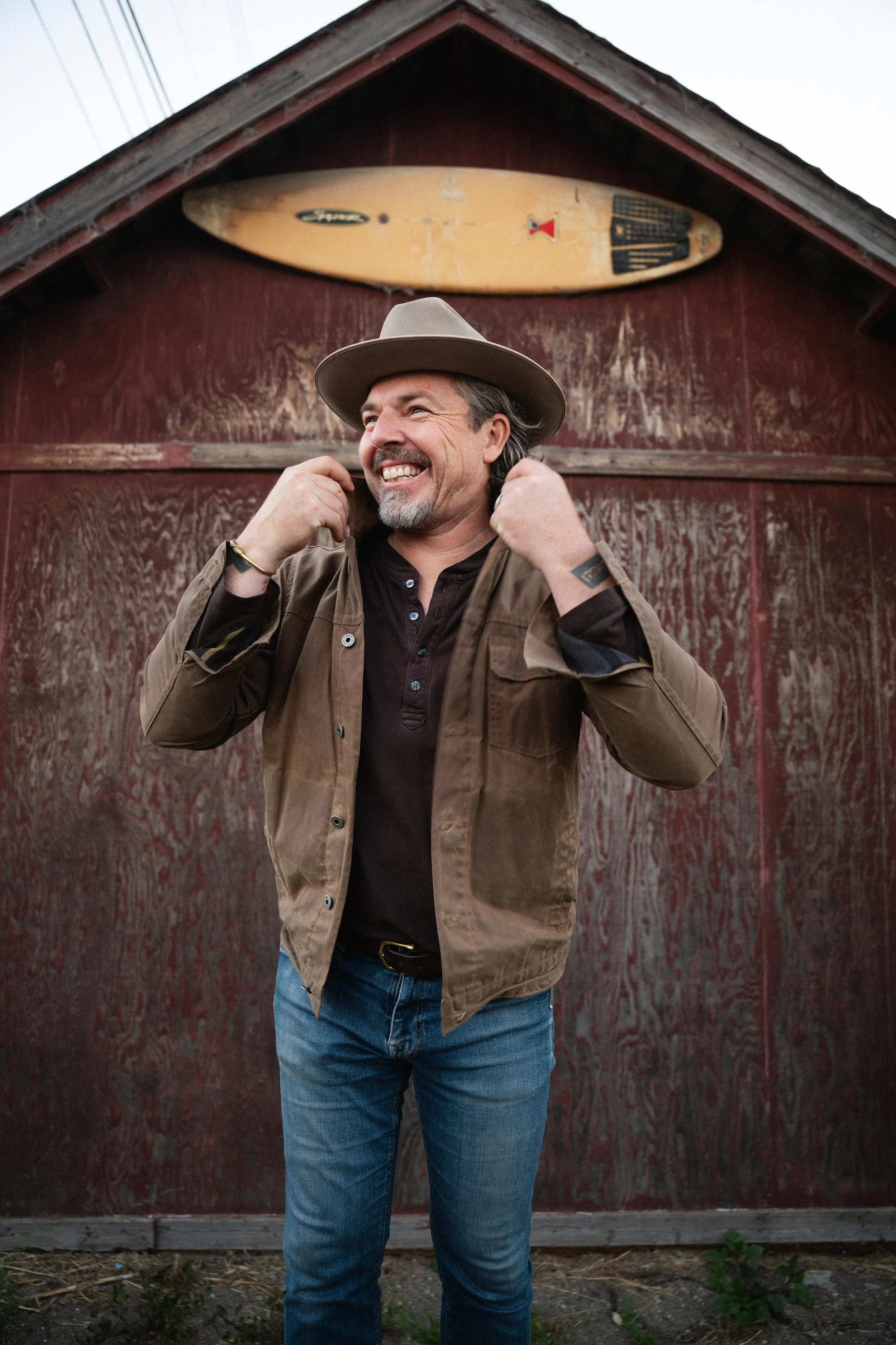 A man with gray hair and a beard wearing a tan wide-brimmed hat and a brown jacket, standing in front of a red wooden barn with a vintage surfboard mounted on it. The man is smiling and adjusting his hat.