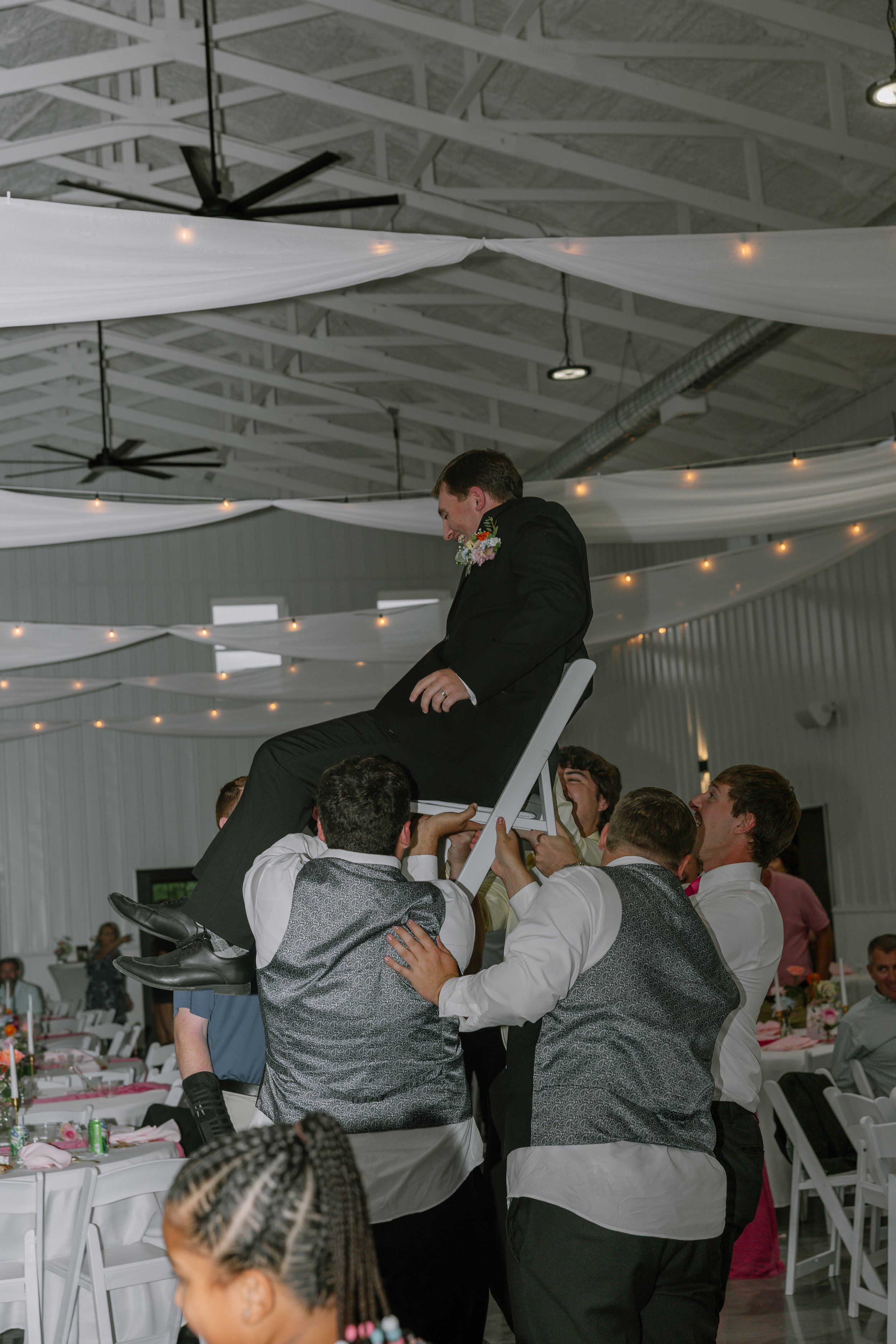 A group of men dressed in formal attire with matching vests perform a chair lift at a wedding reception held indoors with white drapery and string lights hanging from the ceiling.
