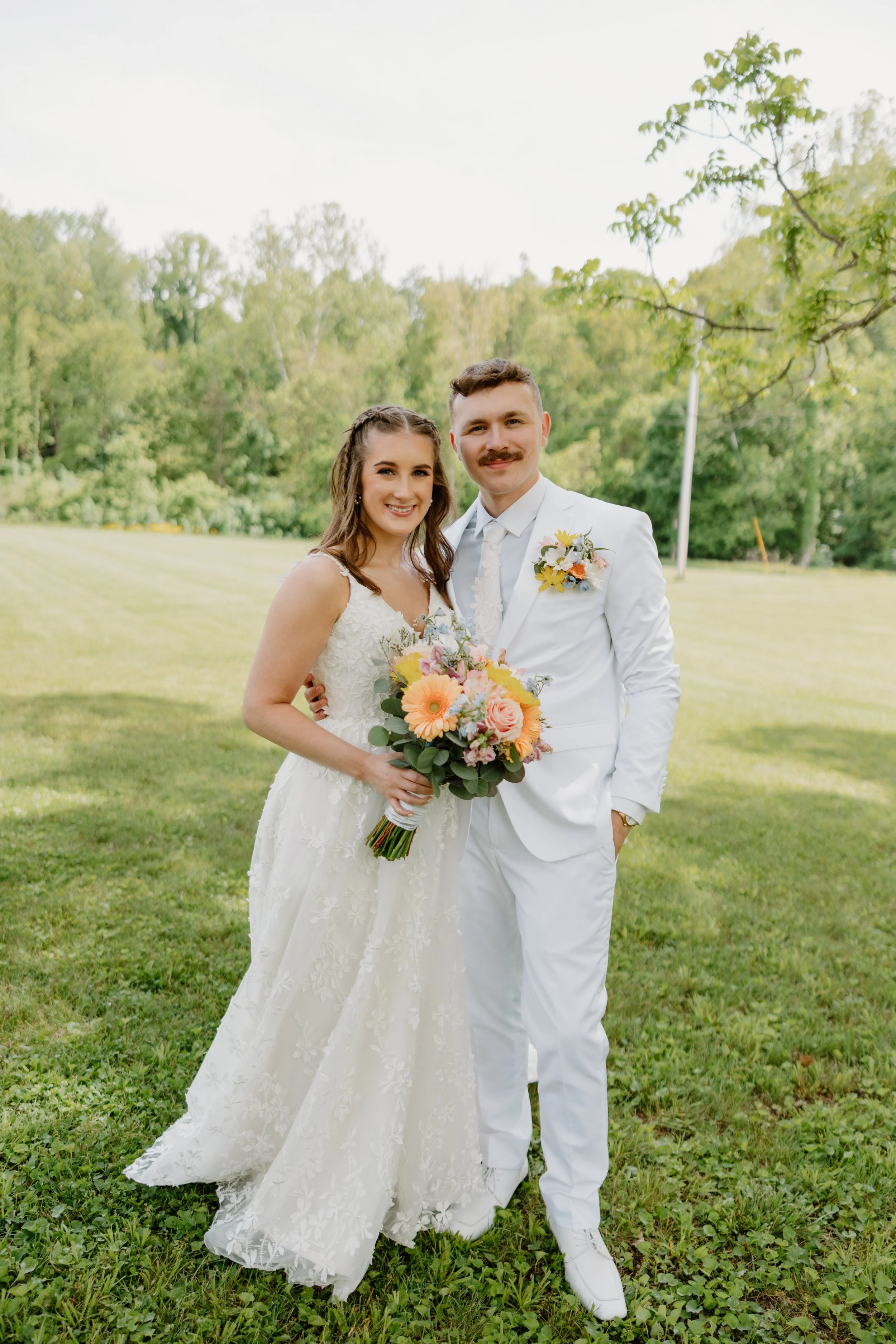 A newlywed couple stands outdoors on a grassy field, with a backdrop of green trees. The bride is holding a colorful bouquet and wearing a white lace wedding dress, while the groom is dressed in an all-white suit with a boutonniere. Both are smiling and facing the camera.