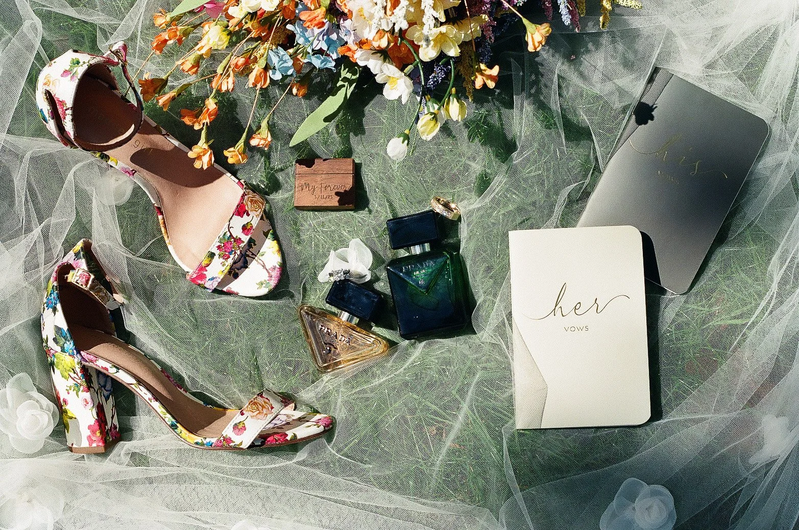A flat lay of women's accessories and items on a sheer, light-colored fabric background. It includes a pair of floral high-heeled shoes, a small jewelry box, two Prada perfume bottles, a mirror, a white card with gold cursive writing that says 'her vows,' and a floral bouquet in the top center.