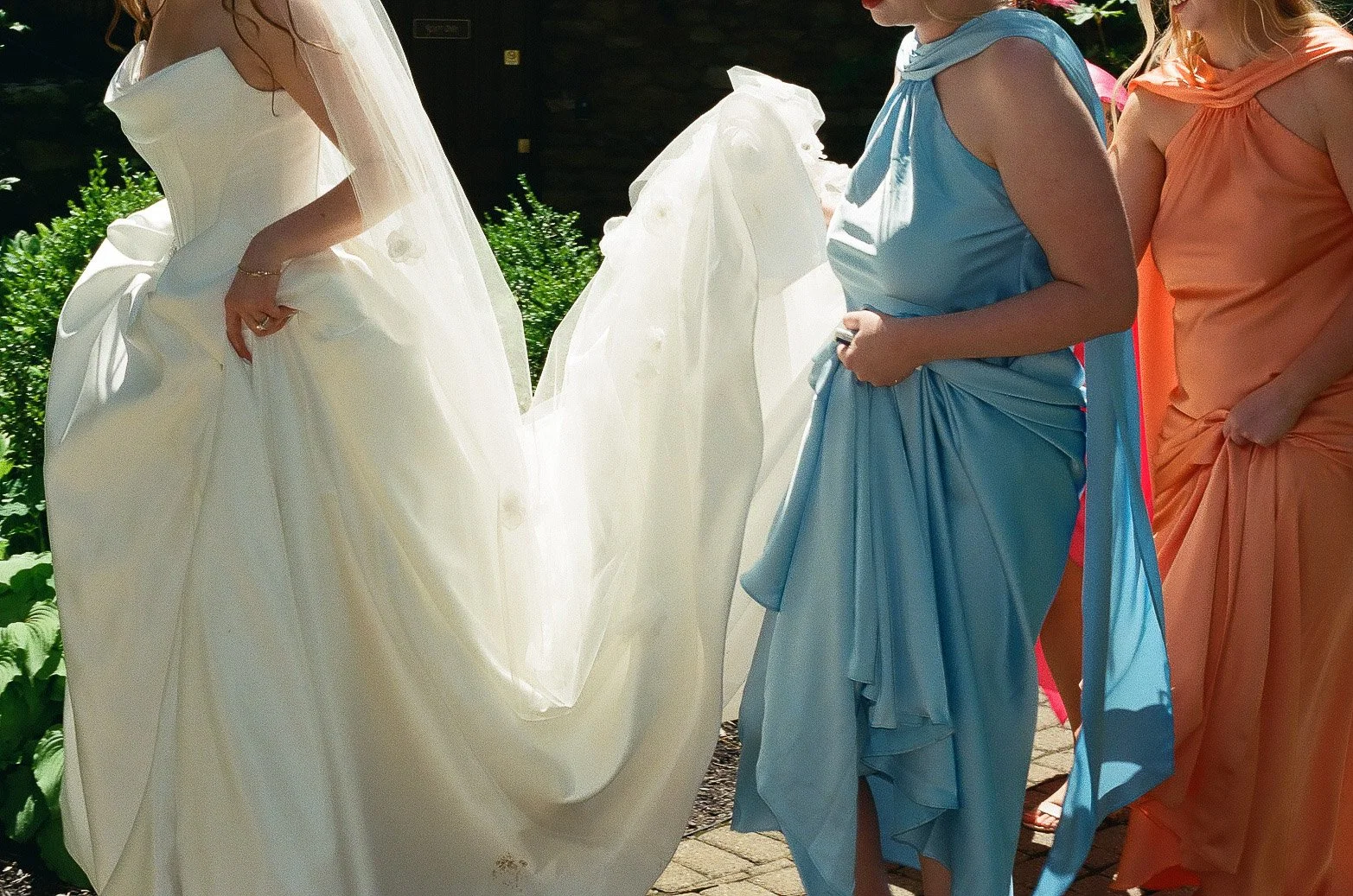 People dressed in colorful formal dresses, with one woman in a white wedding gown and veil, standing outdoors.