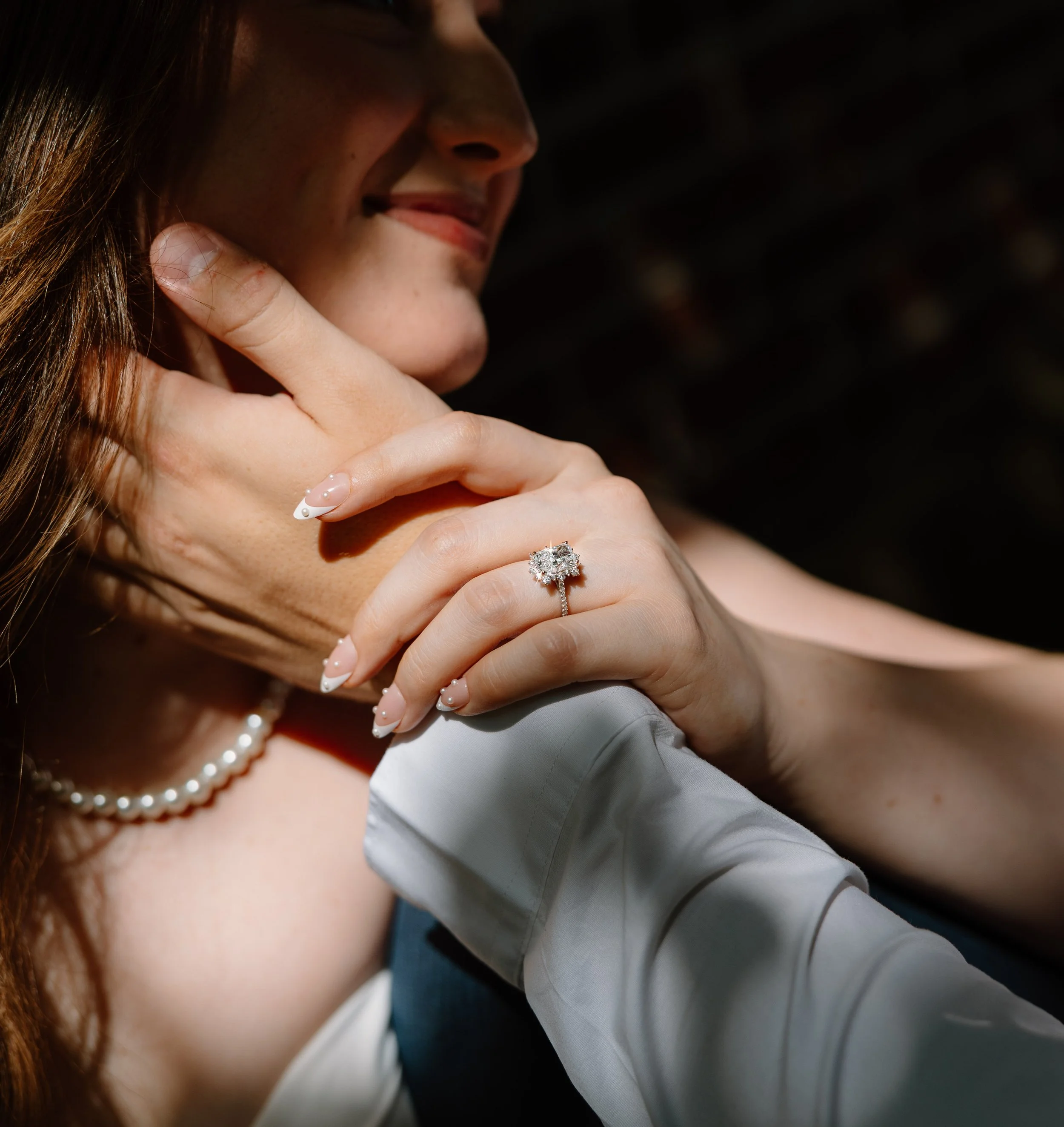Close-up of a woman smiling, showing her teeth, with part of her hand and a diamond ring visible near her chin. She has long, reddish-brown hair and is wearing a pearl necklace. The background is dark.