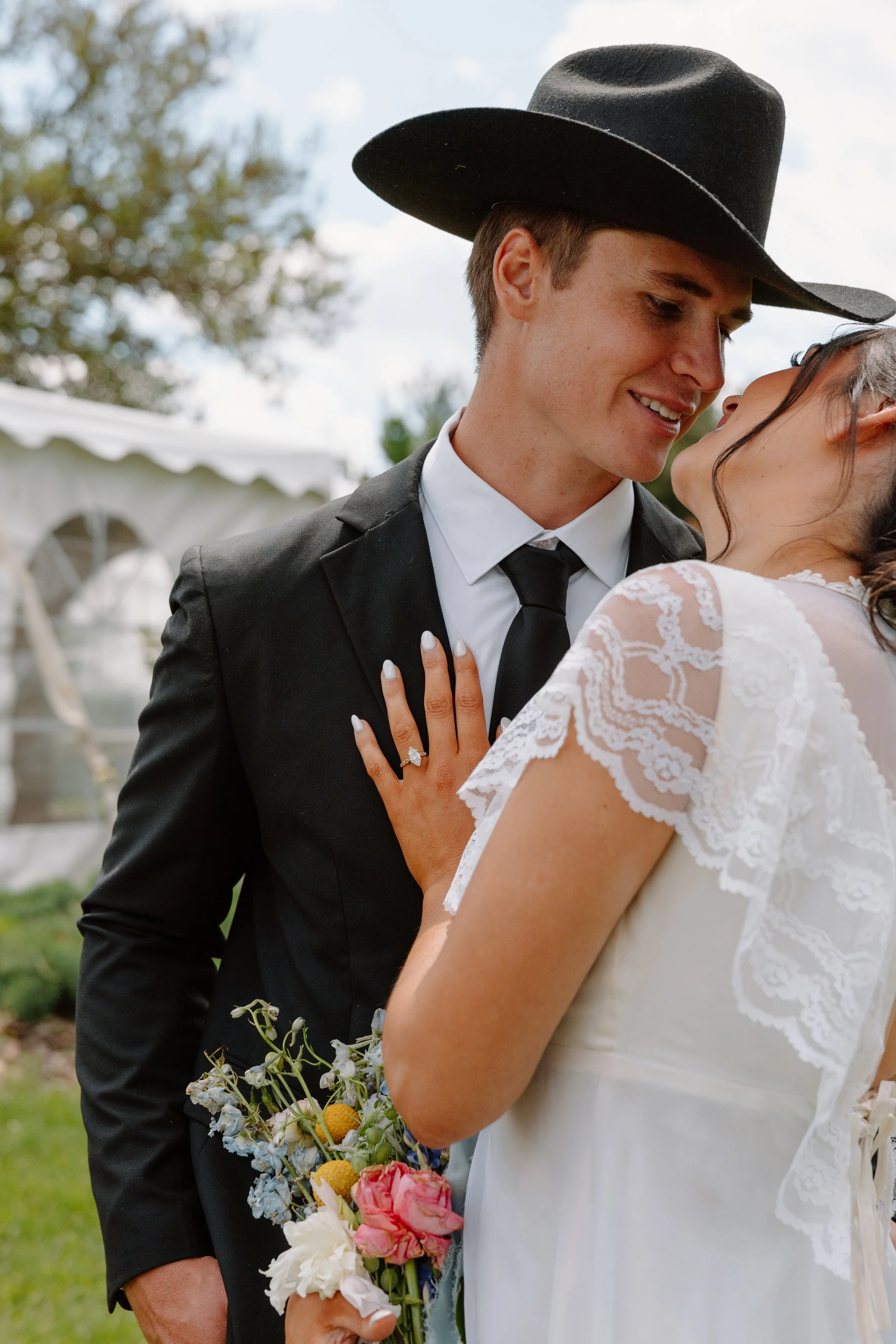 A young man wearing a black suit and black hat is smiling at a young woman in a white lace dress, with both leaning close to each other outdoors on a bright day. The woman is holding a colorful bouquet of flowers.