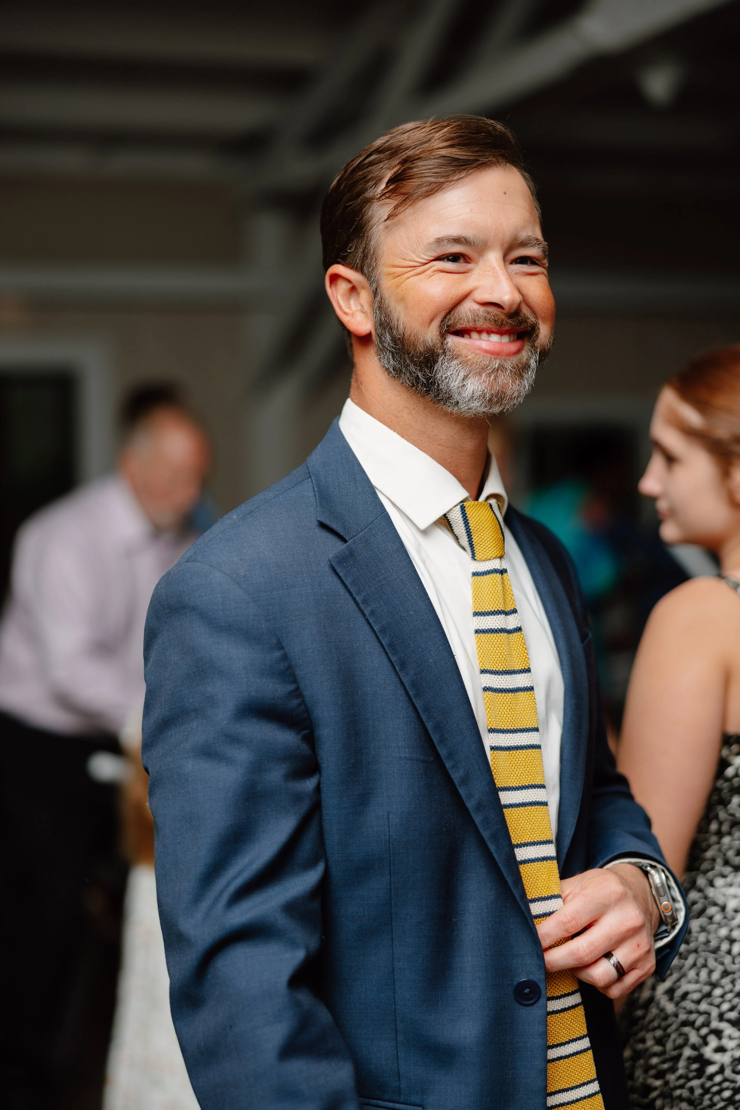 Smiling man with beard and brown hair wearing a blue suit, striped yellow and blue tie, and silver watch, at a social event with blurred people in the background.