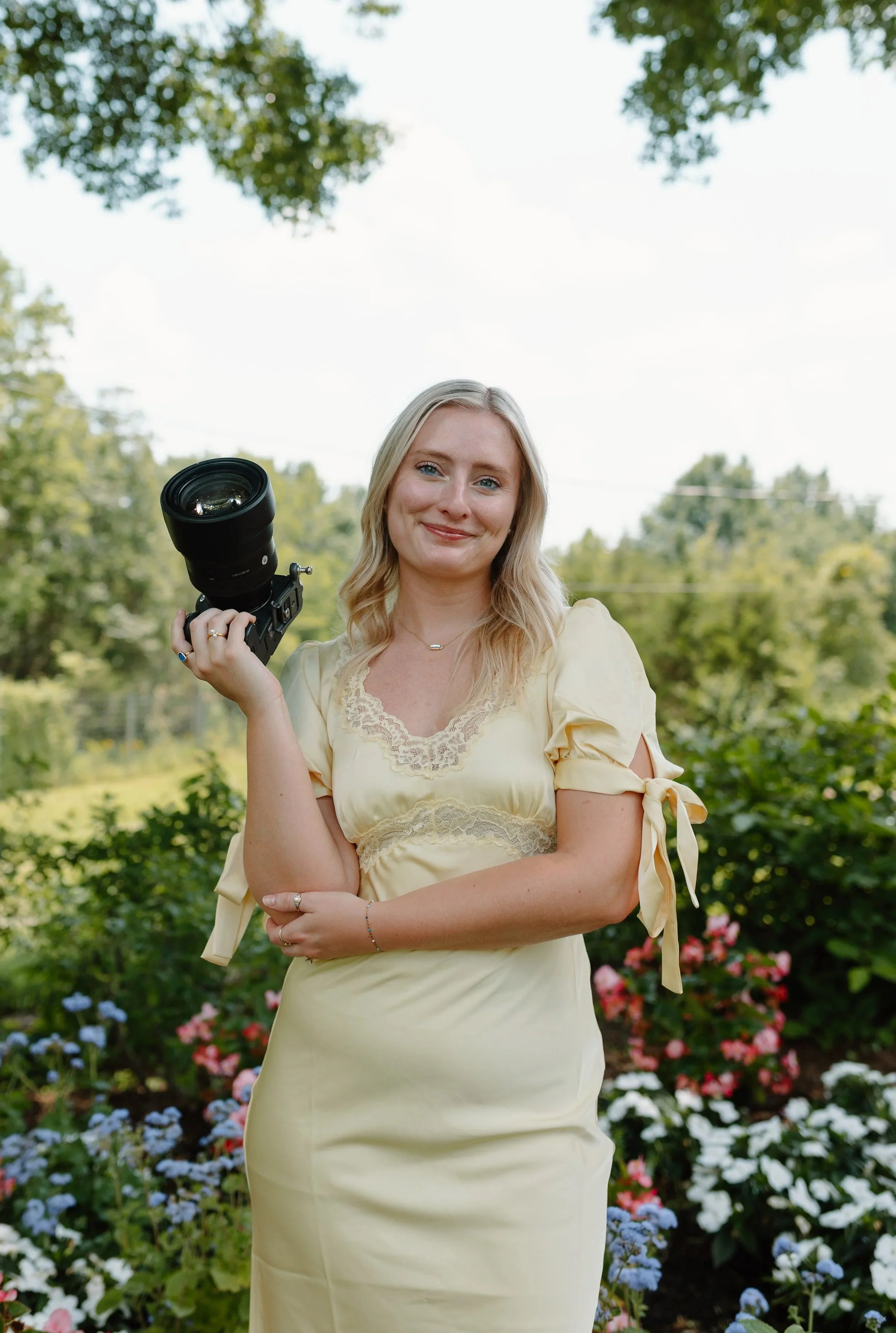 A woman in a yellow dress holding a camera standing outdoors in a garden with colorful flowers and greenery.