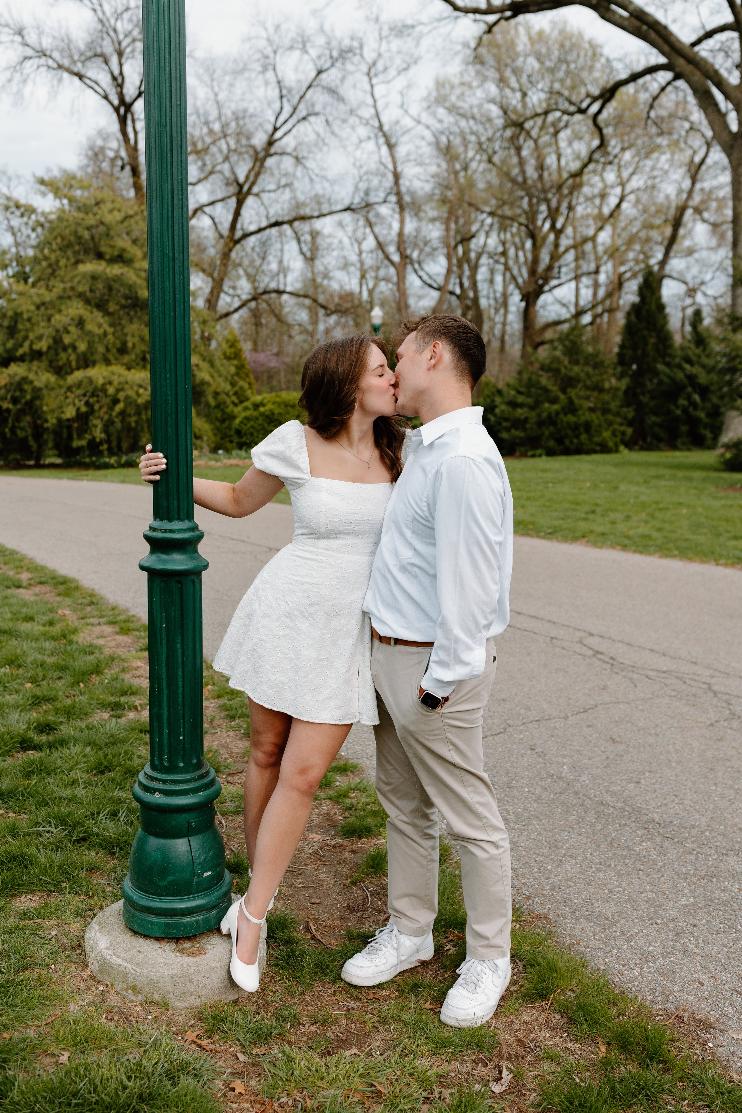 A couple sharing a kiss outdoors, with the woman wearing a white dress and high heels, and the man dressed in beige pants and a white shirt, standing next to a green park lamp post in a park with trees and grass.