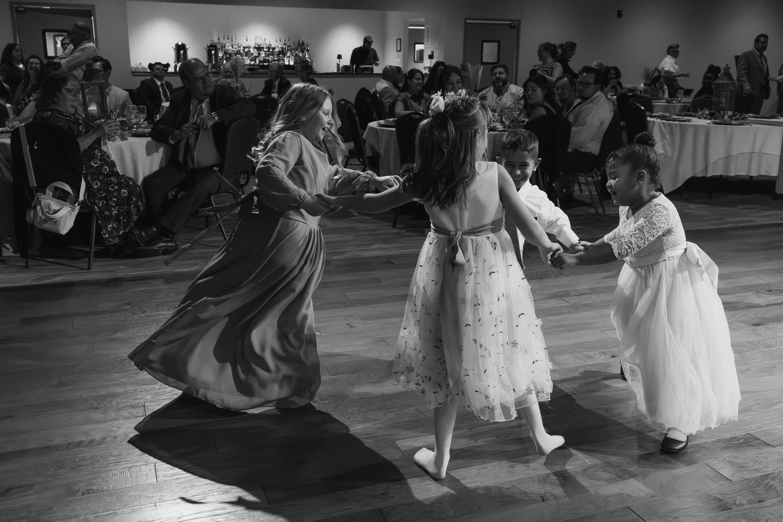 Four young girls in dresses are holding hands and dancing in a circle at a social event, while guests sit at tables watching in the background.