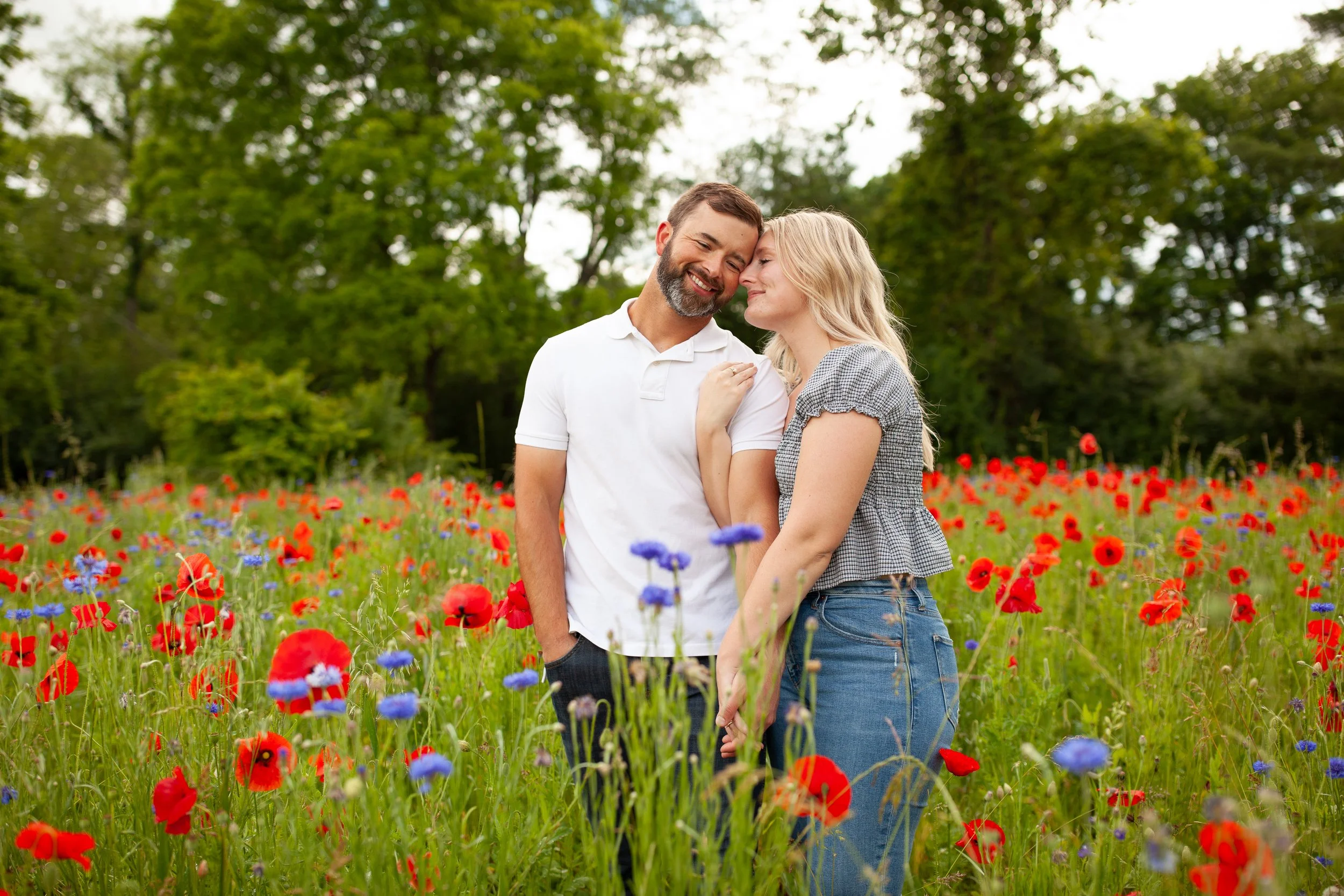A man and woman smiling and leaning their foreheads together in a field of red and blue flowers with green trees in the background.