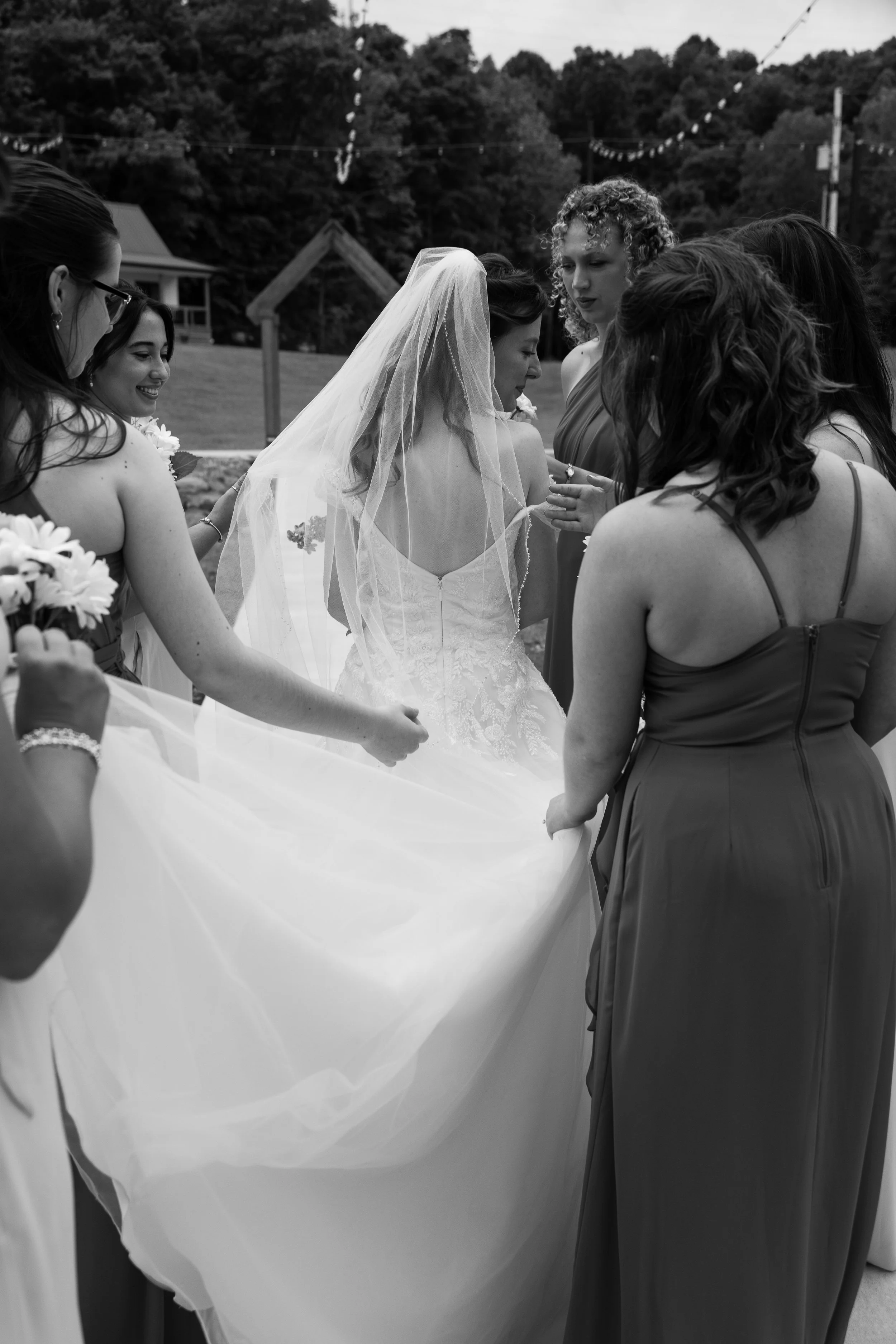 A bride in a wedding dress and veil surrounded by her bridesmaids, holding her gown's train in an outdoor setting with trees and string lights in the background.
