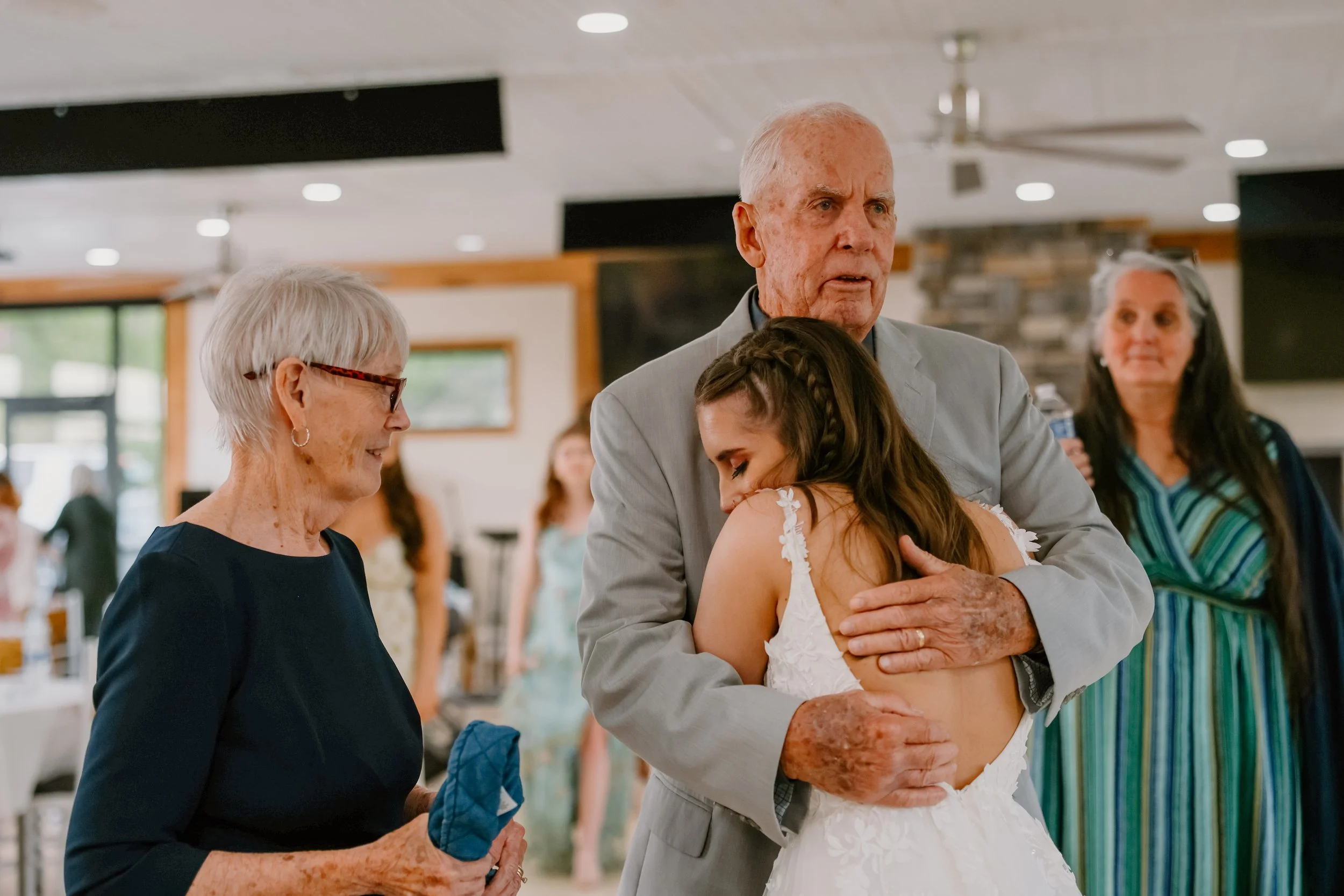 An elderly man in a gray suit hugging a young woman in a white wedding dress, with an older woman in a navy dress and glasses looking on, in a decorated indoor celebration setting.