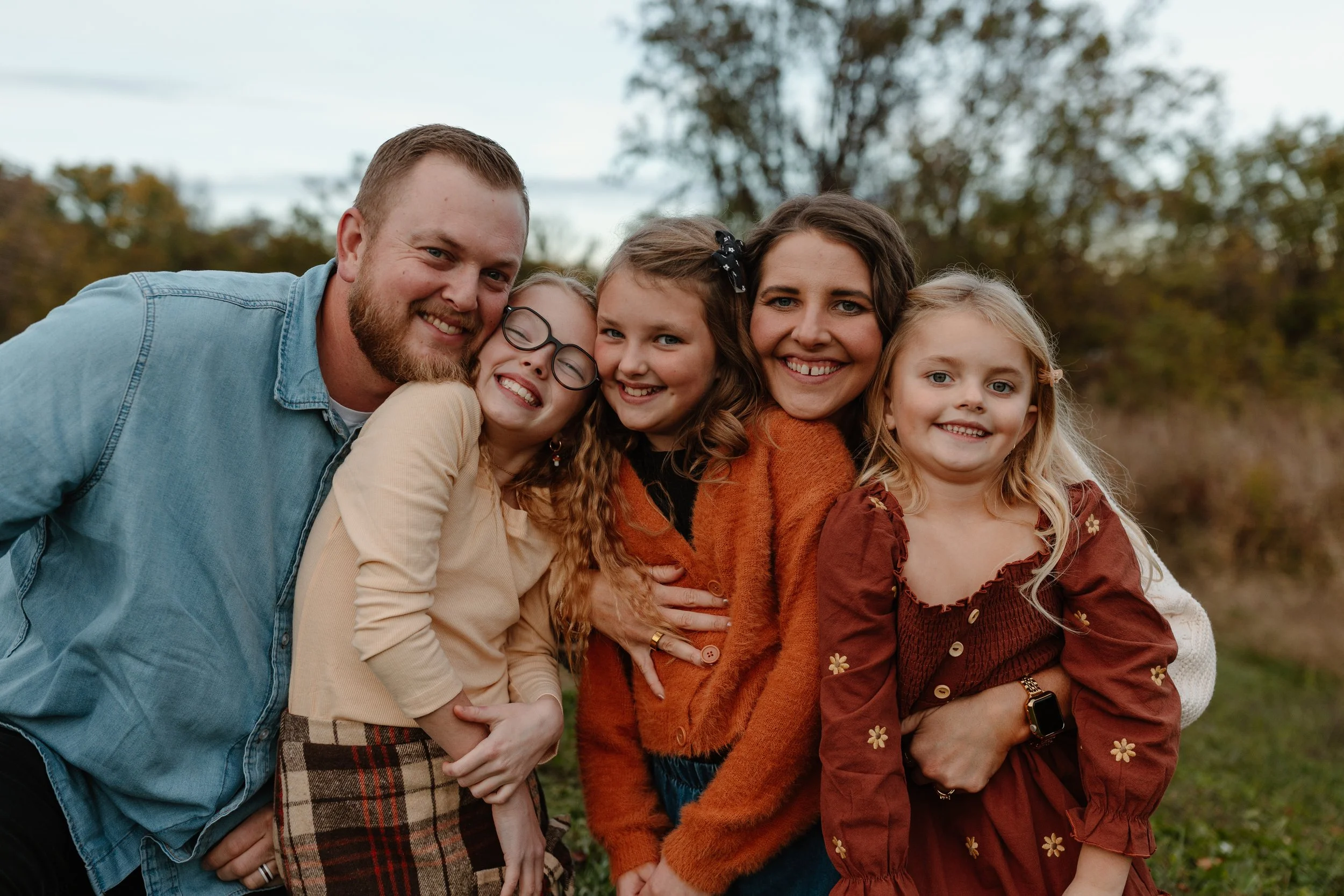 A family of five, including a man, a woman and three young girls, is posing together outdoors with trees in the background. They are smiling and close to each other.