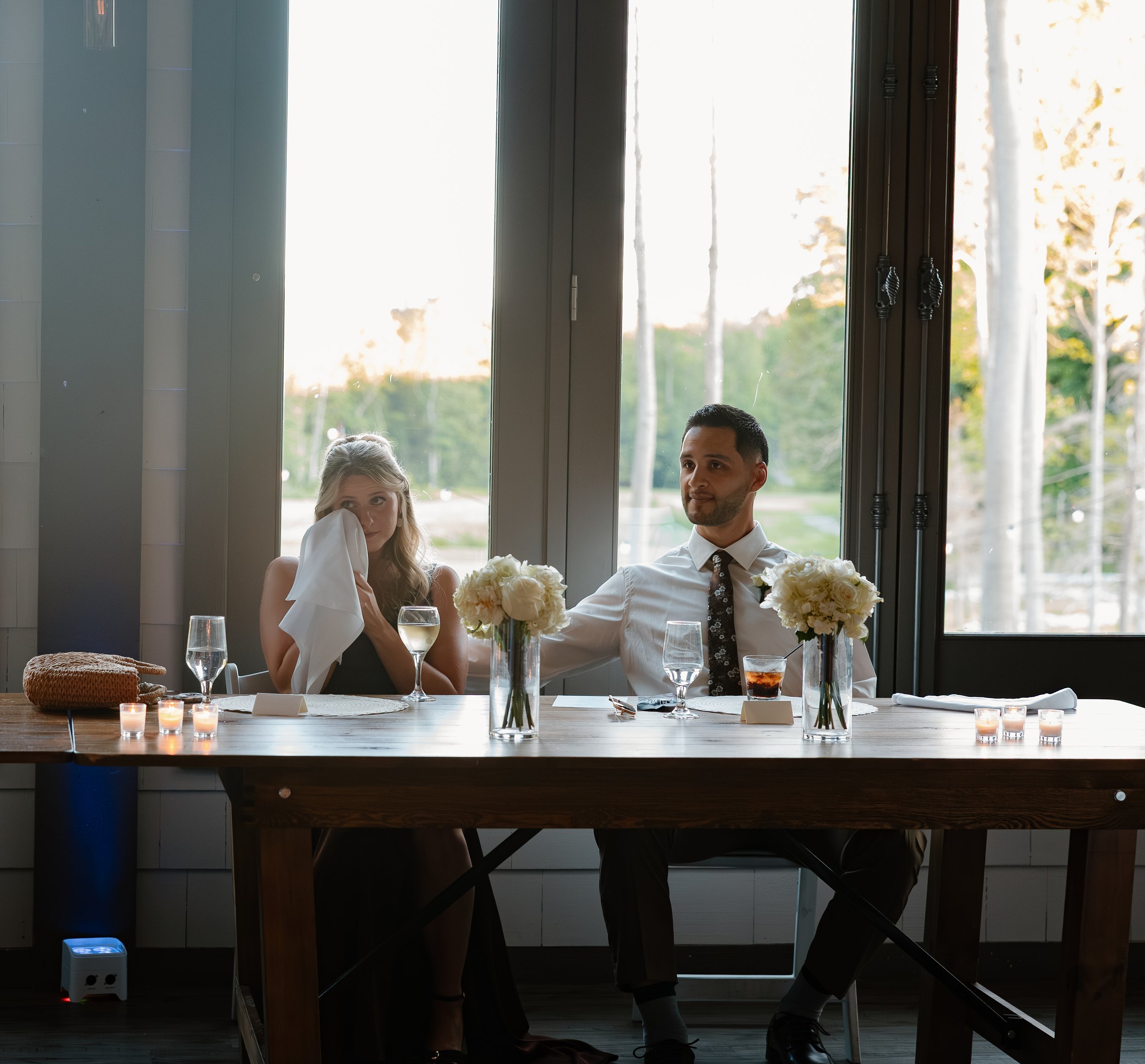 A couple is sitting at a dining table having a meal. The woman is wiping her face with a napkin, and the man is looking ahead. The table is decorated with white flowers, glasses of water, a beverage, and candles. They are indoors near a large window 