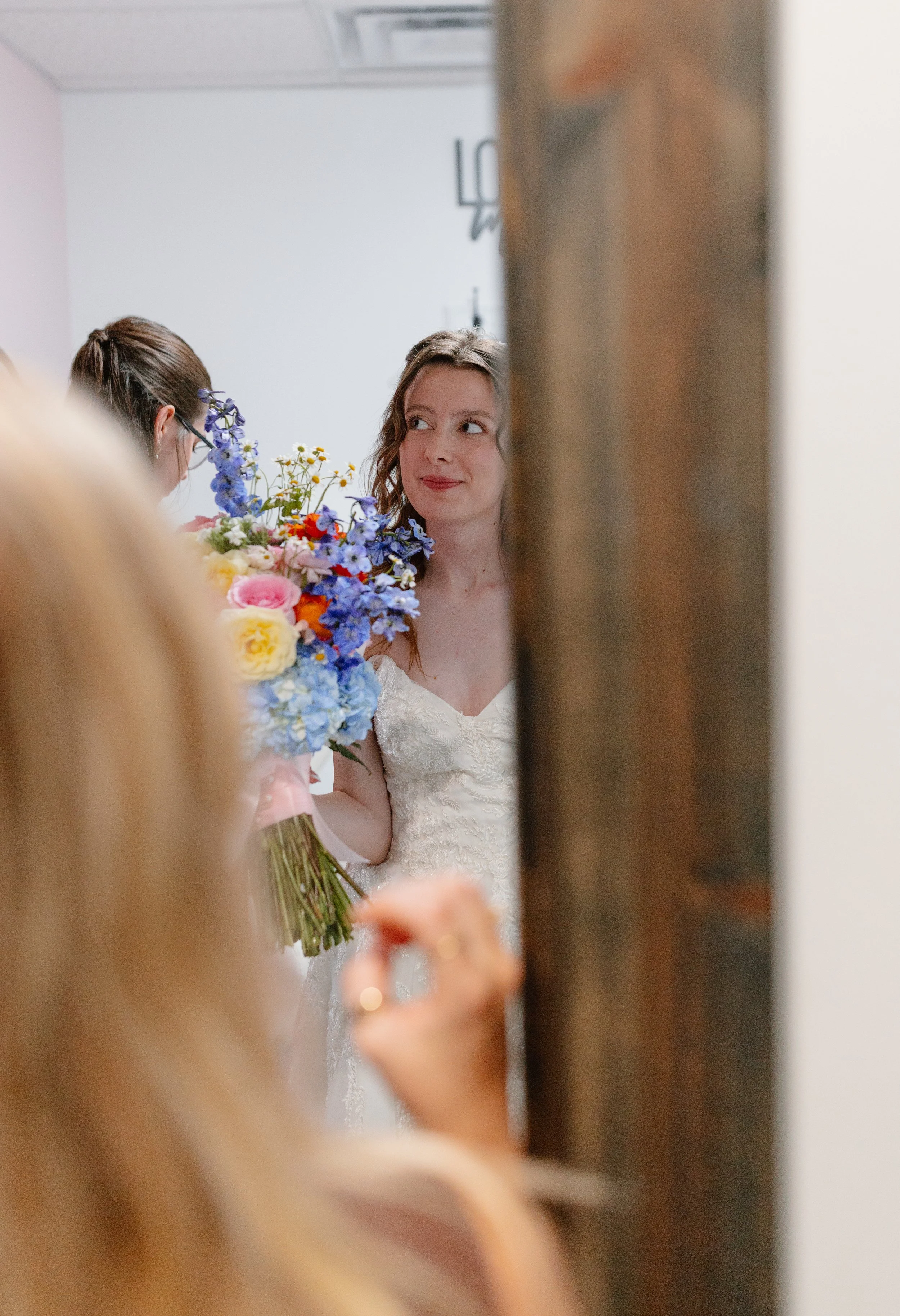 A bride with light brown, wavy hair, wearing a white lace wedding dress, holding a colorful bouquet, and smiling softly while looking at her reflection in a mirror, with a blurred person holding a flower in the foreground.