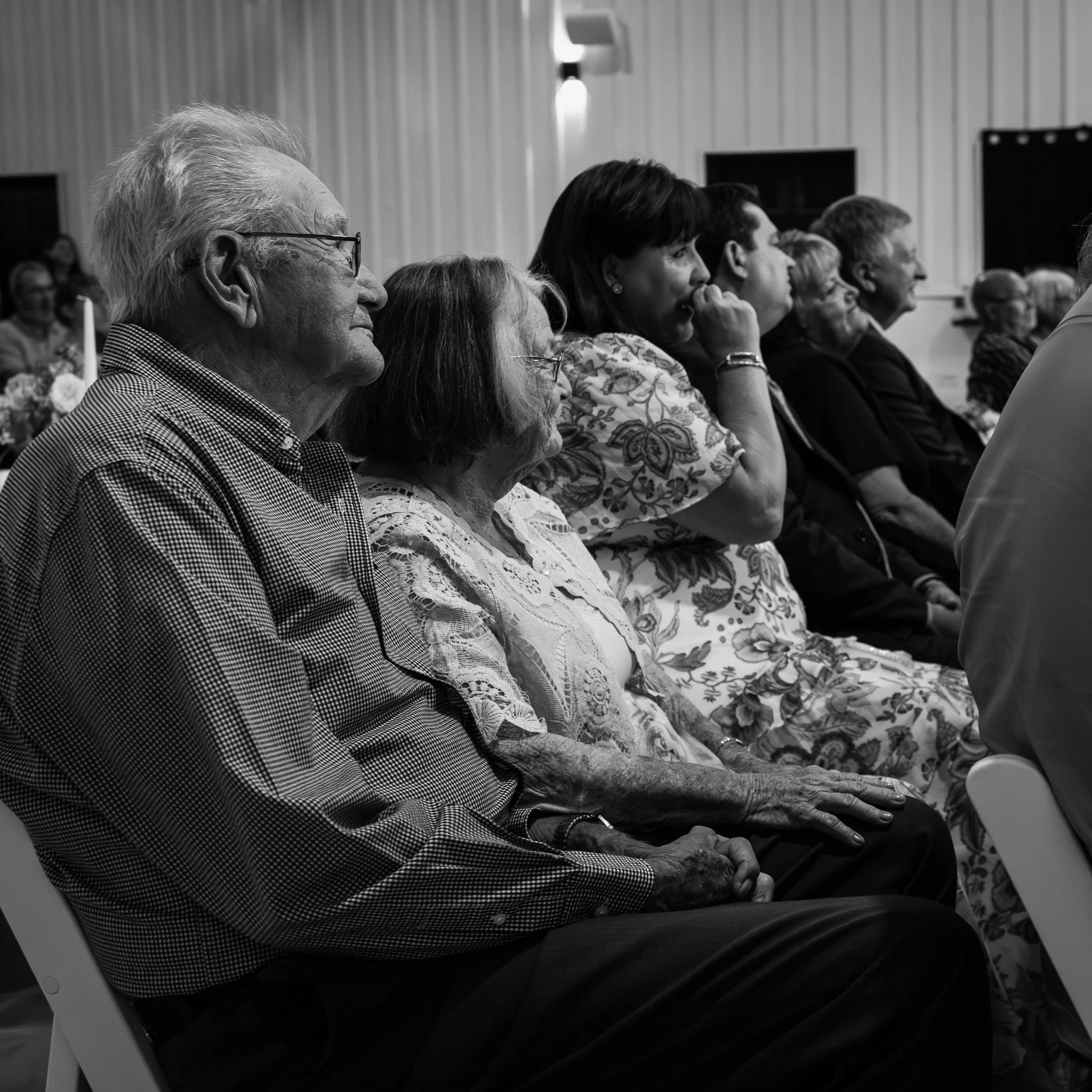 A black-and-white photo of elderly people sitting in a row, attentively listening at an indoor event. The image shows diverse individuals, some with glasses, dressed in casual and semi-formal clothing, with a plain wall and a wall lamp in the background.
