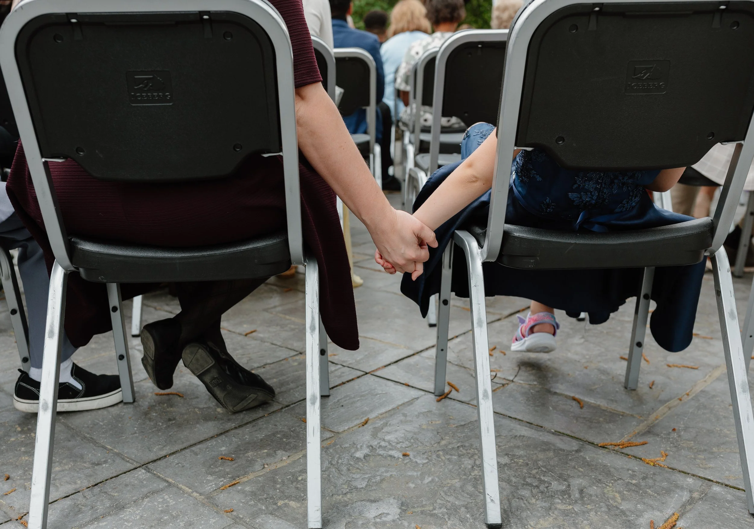 People attending an outdoor event sitting on chairs, with two individuals in the foreground holding hands underneath the chairs, one wearing a maroon top and the other a blue dress. The scene is on a paved surface with some fallen leaves.