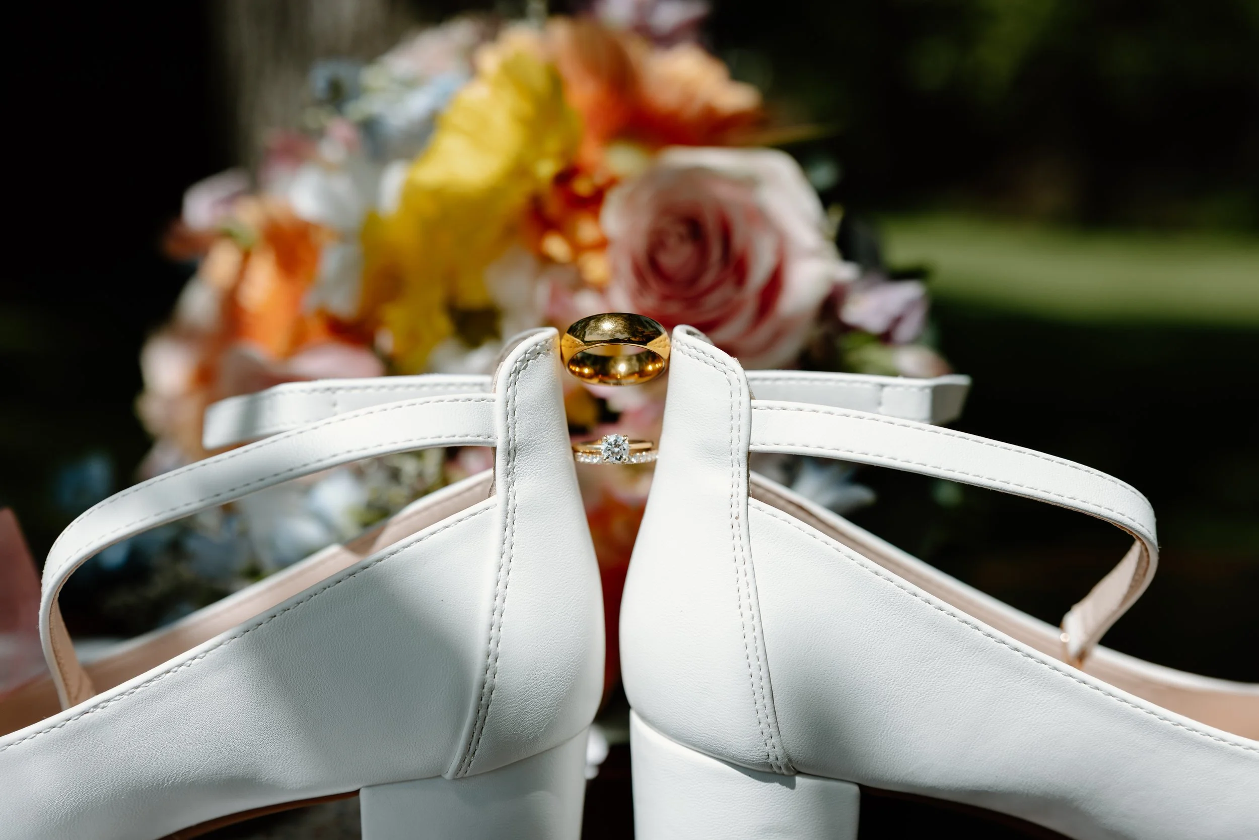 A pair of white high-heeled shoes with a pair of gold rings placed on top, with a blurred bouquet of flowers in the background.