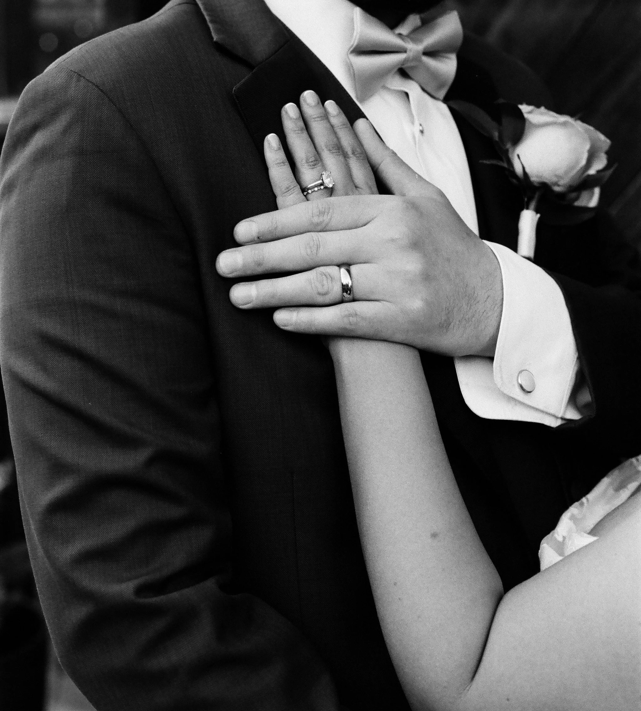 Black and white photo of a groom in a tuxedo with a bow tie, and a bride with her hand on his shoulder showing her wedding ring, including an engagement ring with a large stone. The bride is wearing a white dress with a cuffed sleeve. The groom's boutonniere is a rose.