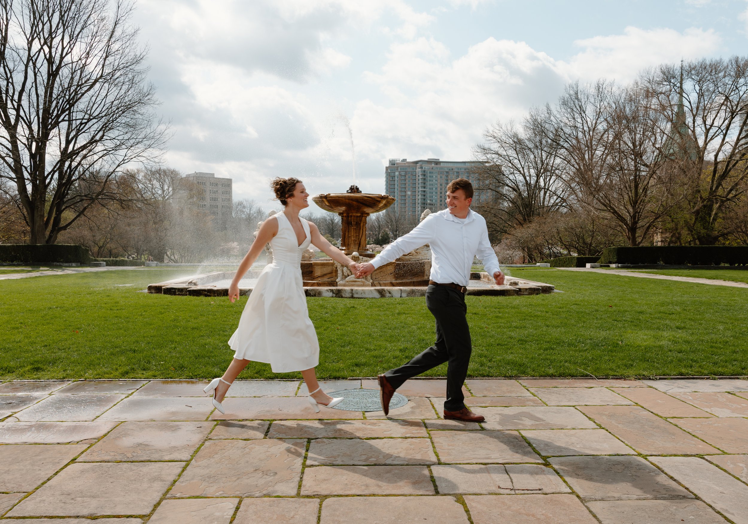 A newlywed couple holding hands and dancing outdoors in a park with a stone fountain, trees, and high-rise buildings in the background.