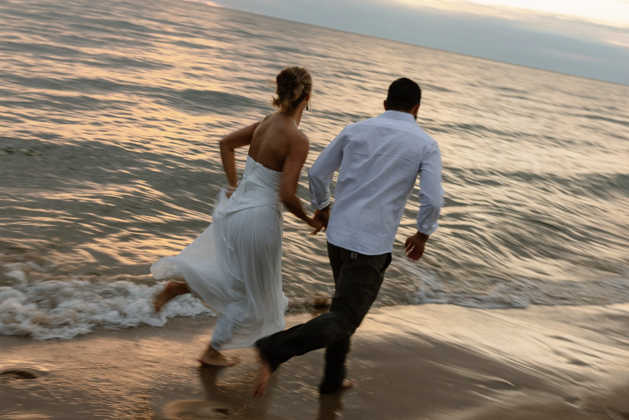 A man and a woman holding hands and running along the shoreline at sunset, with the woman wearing a white dress and the man dressed in a white shirt and dark pants.