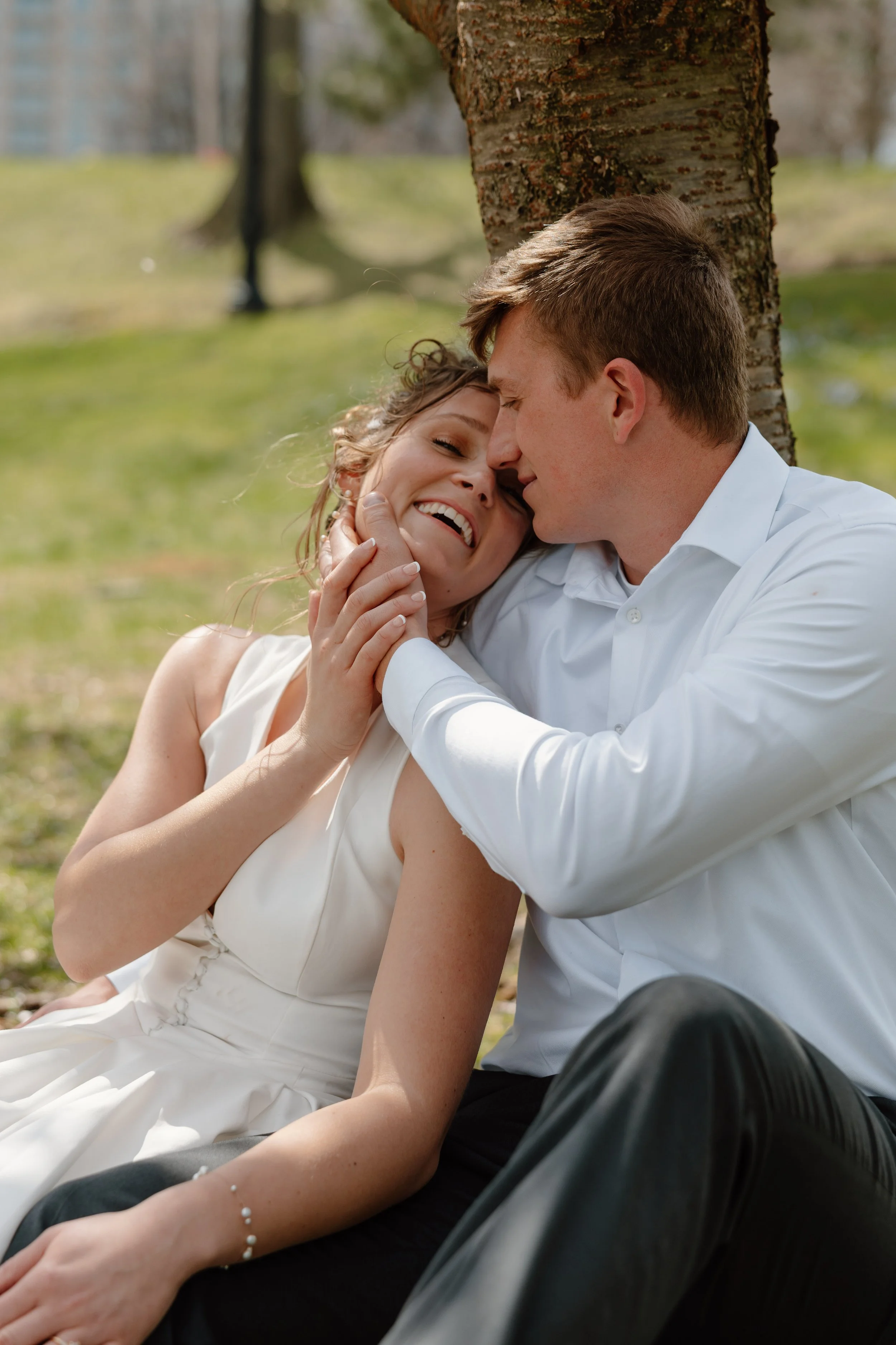A couple dressed in wedding attire sitting outdoors under a tree, sharing a moment of affection with their foreheads touching. The woman is smiling with her eyes closed, and the man is gently holding her face. The background is a park-like setting with blurred trees and grass.