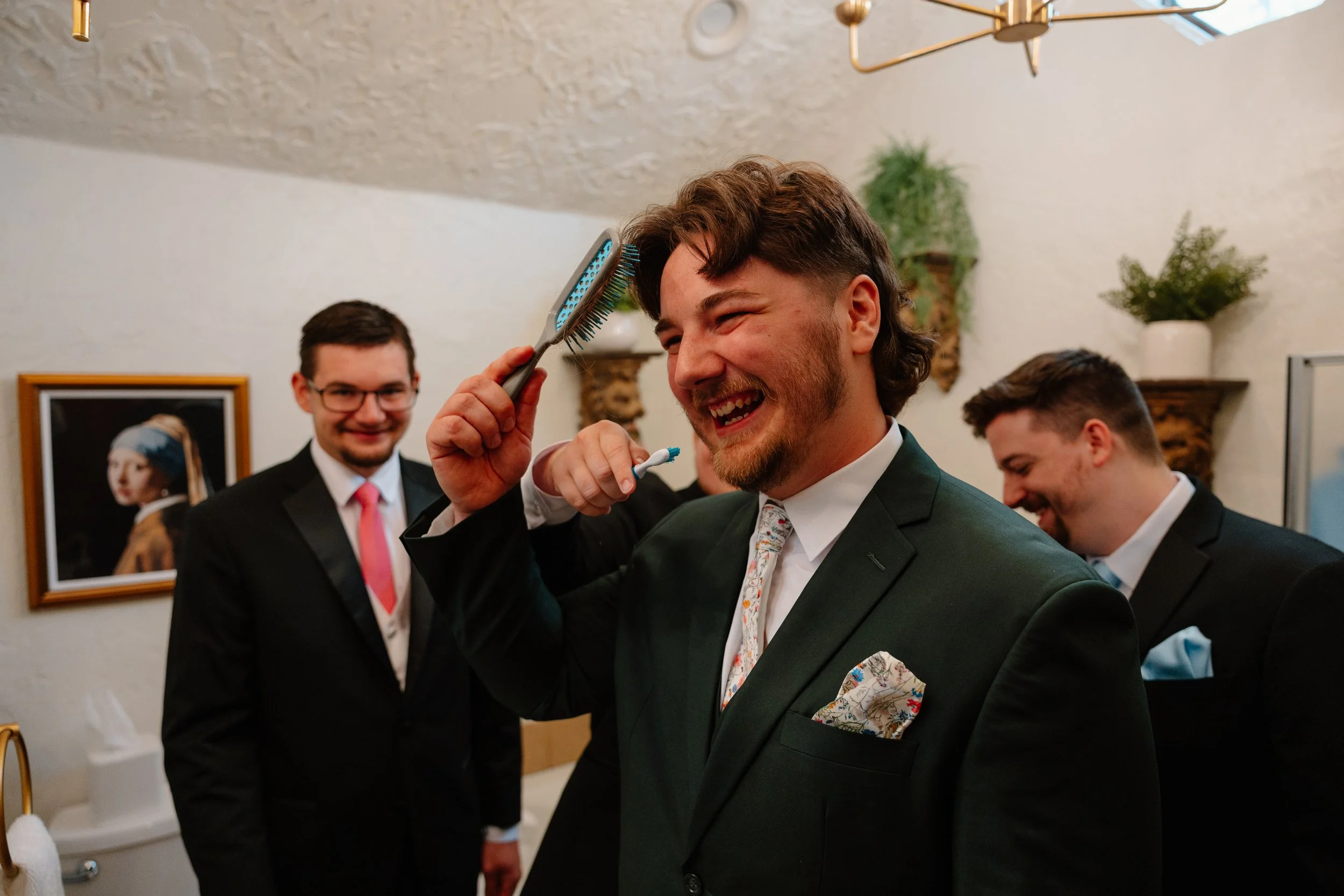 A group of men dressed in formal suits, smiling and having fun at an indoor gathering. One man is brushing another man's hair with a hairbrush, while others look on and smile.
