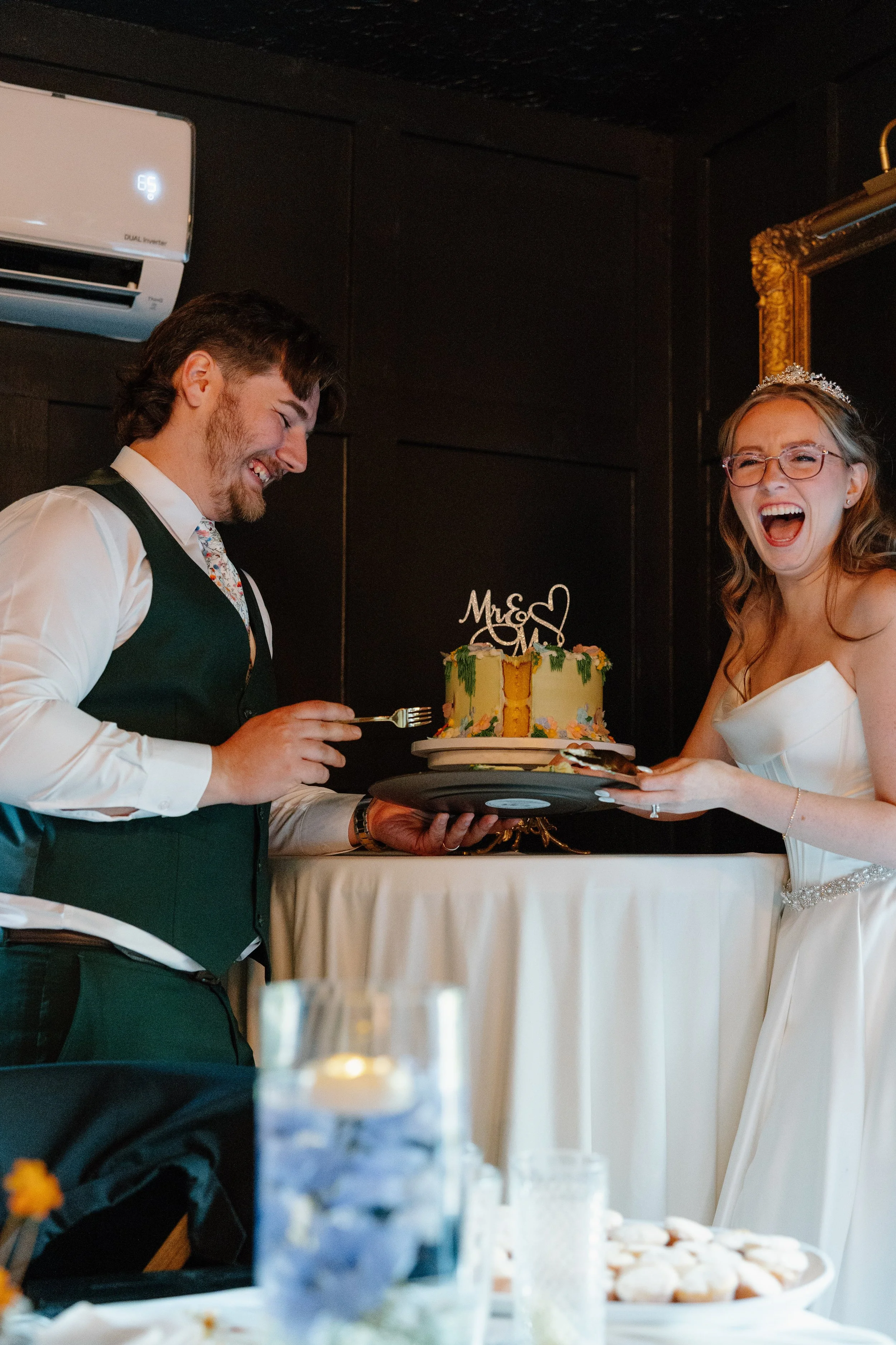 A bride and groom laughing during their wedding cake cutting ceremony. The bride is wearing a white wedding dress and tiara, and the groom is dressed in a dark vest and tie. The wedding cake is decorated with colorful flowers and has a 'Mr & Mrs' top