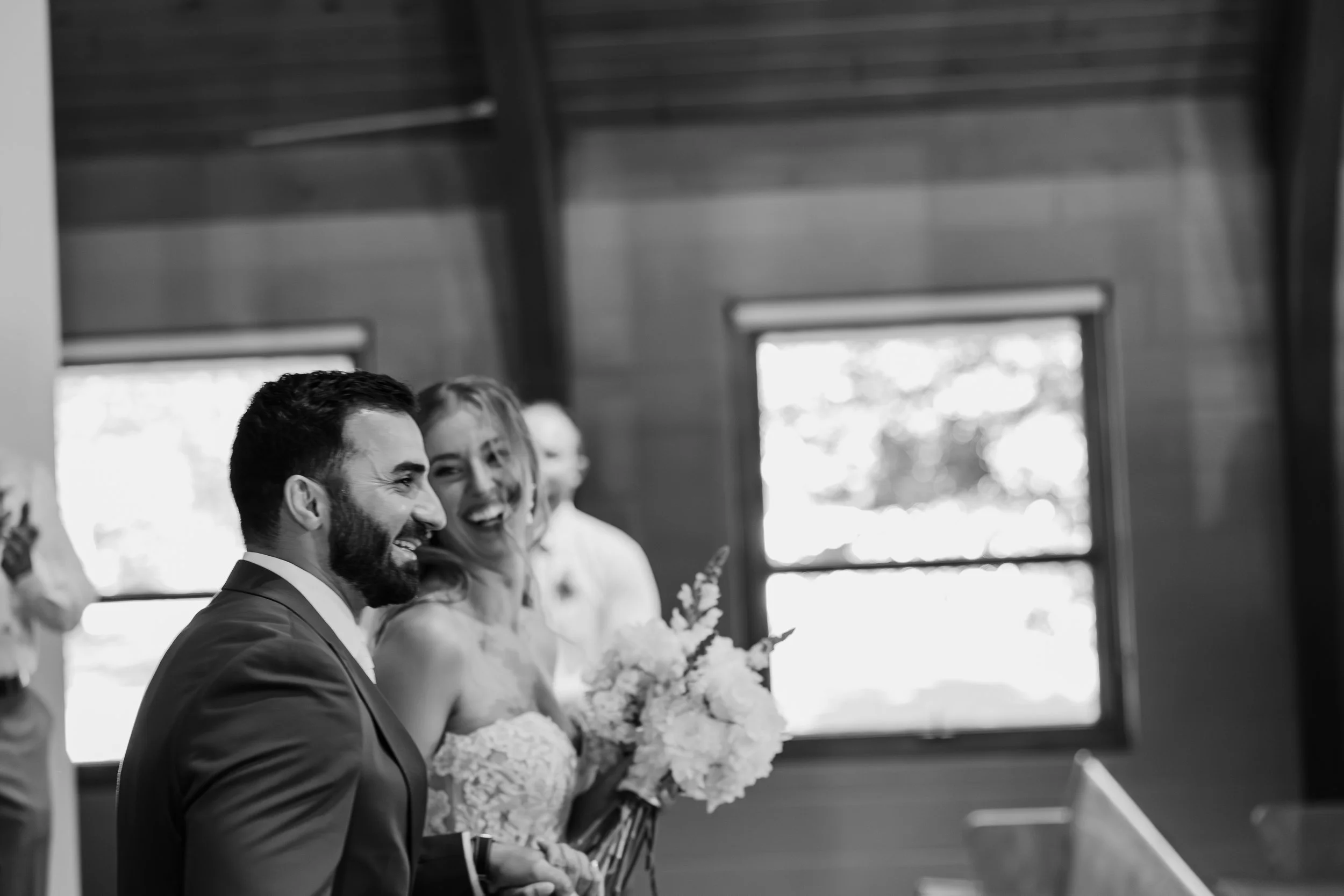 A group of people, including a man in a suit and a woman, smiling and holding bouquets of flowers, gathered in a room with wooden walls and windows in the background.