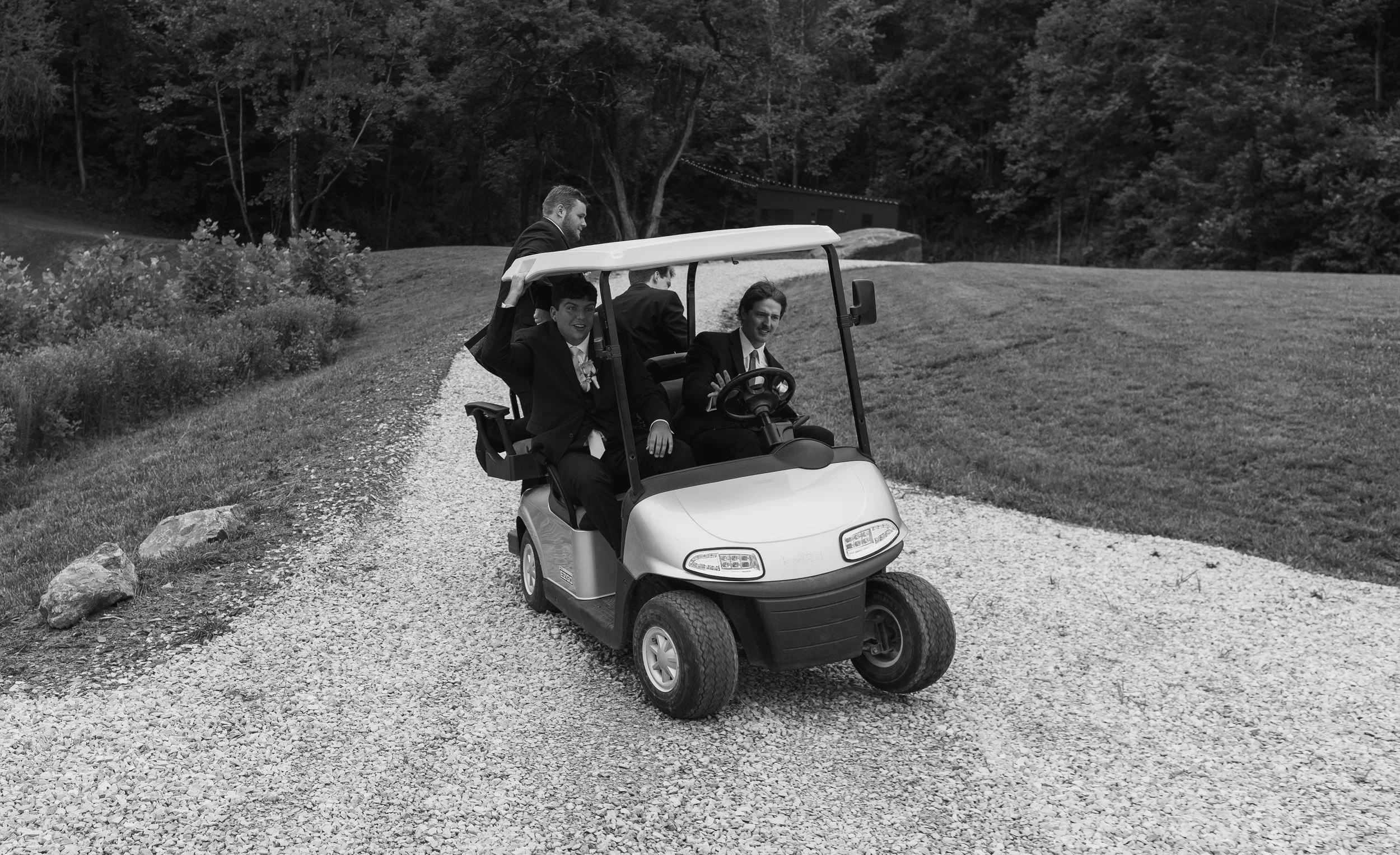 Four men in suits riding in a golf cart on a gravel path in a park or garden area with trees and grass in the background.
