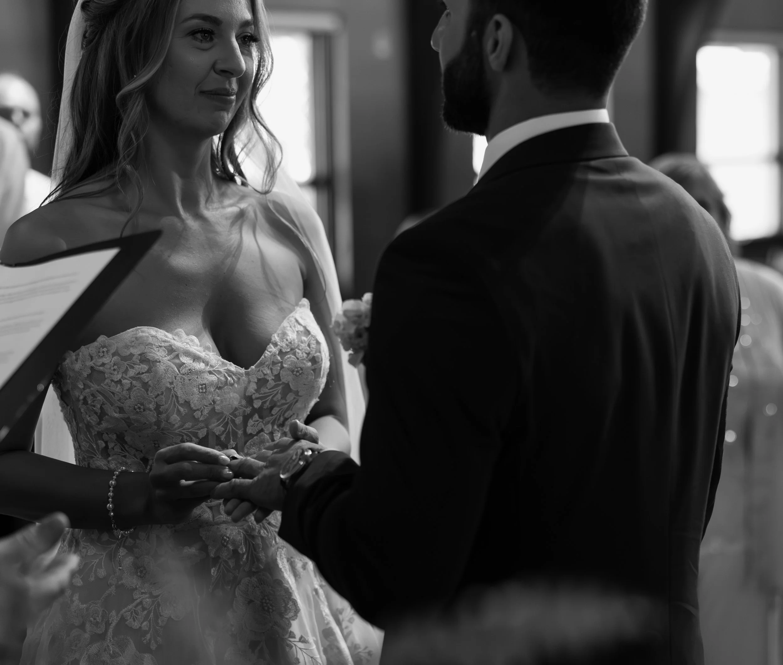 Black and white photo of a bride and groom facing each other during a wedding ceremony, with the bride wearing a lace wedding dress and holding the groom's hands.