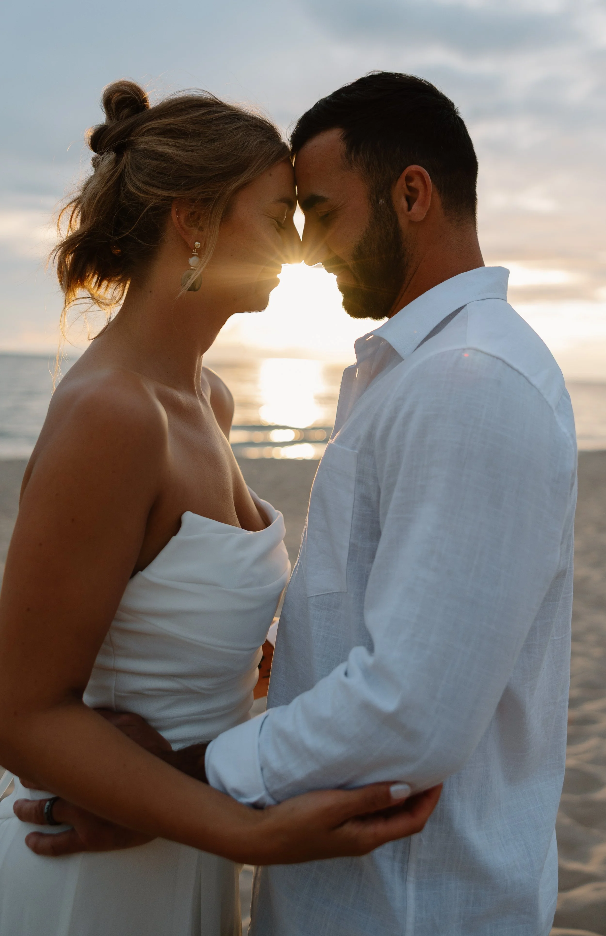 A couple standing close together on a beach during sunset, with their foreheads touching and eyes closed, creating a romantic atmosphere.
