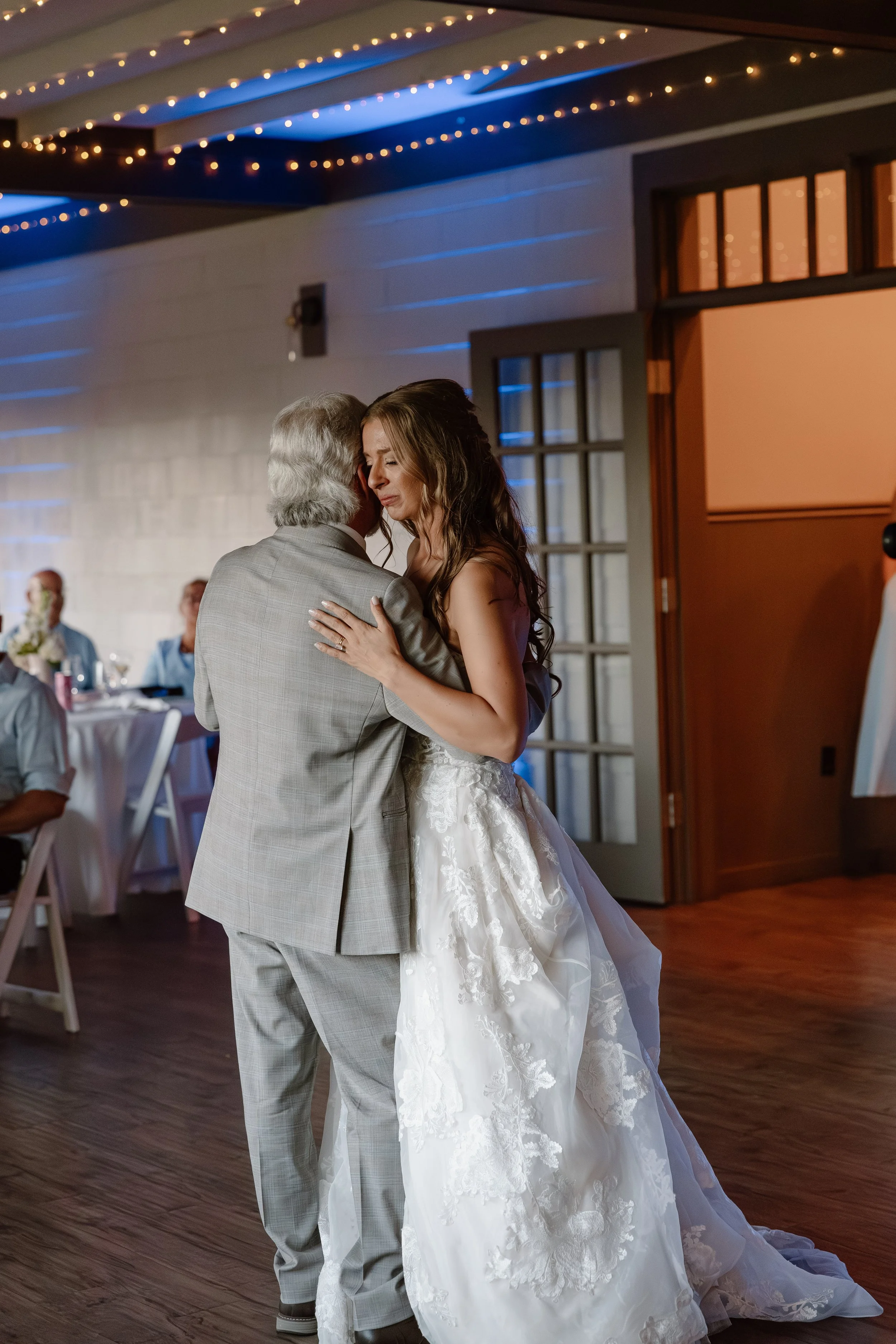 A bride and an older man sharing a dance at a wedding reception, with guests seated at tables in the background and string lights hanging from the ceiling.