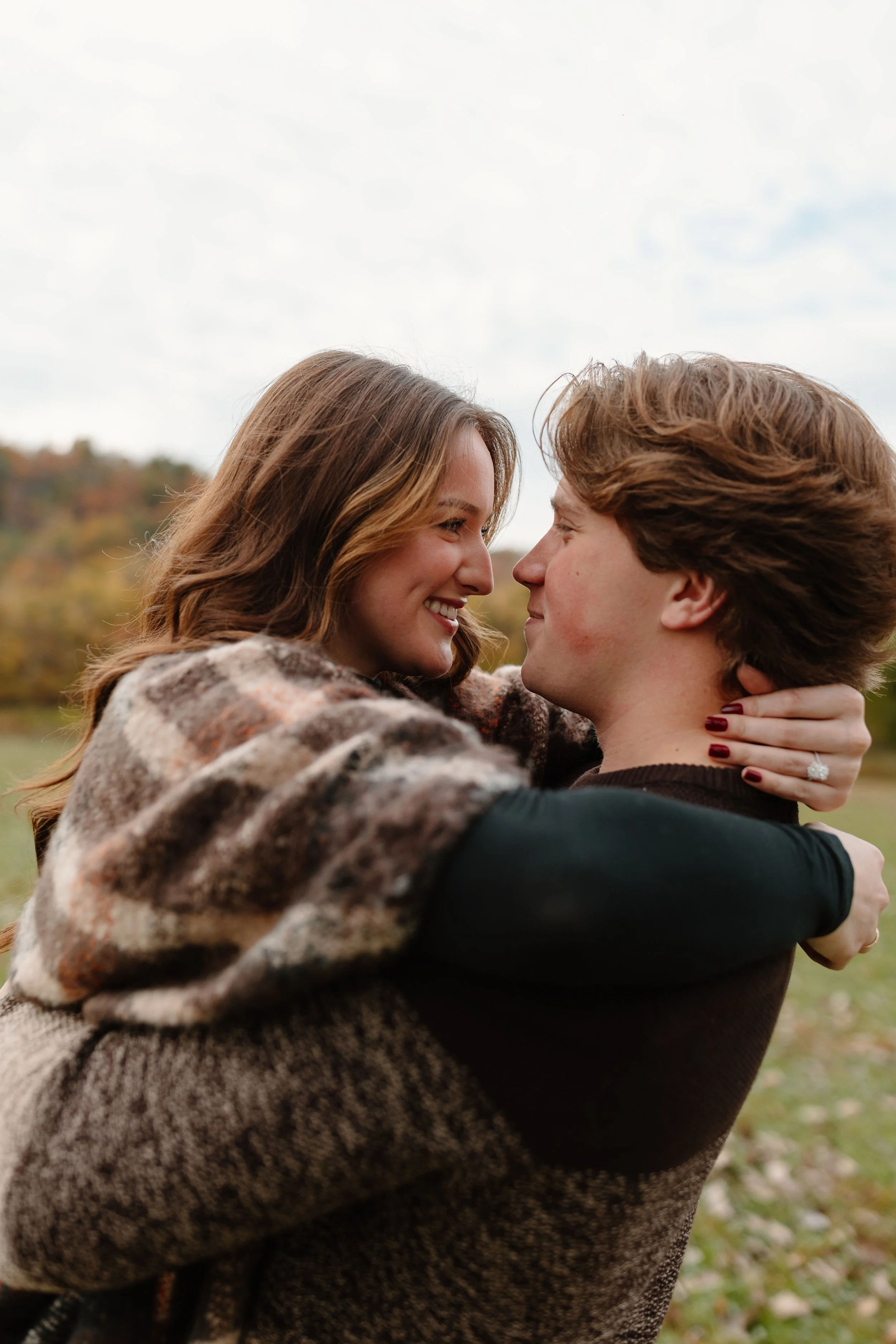 A young woman and a young man hugging outdoors in a park, smiling and gazing into each other's eyes, with trees and cloudy sky in the background.