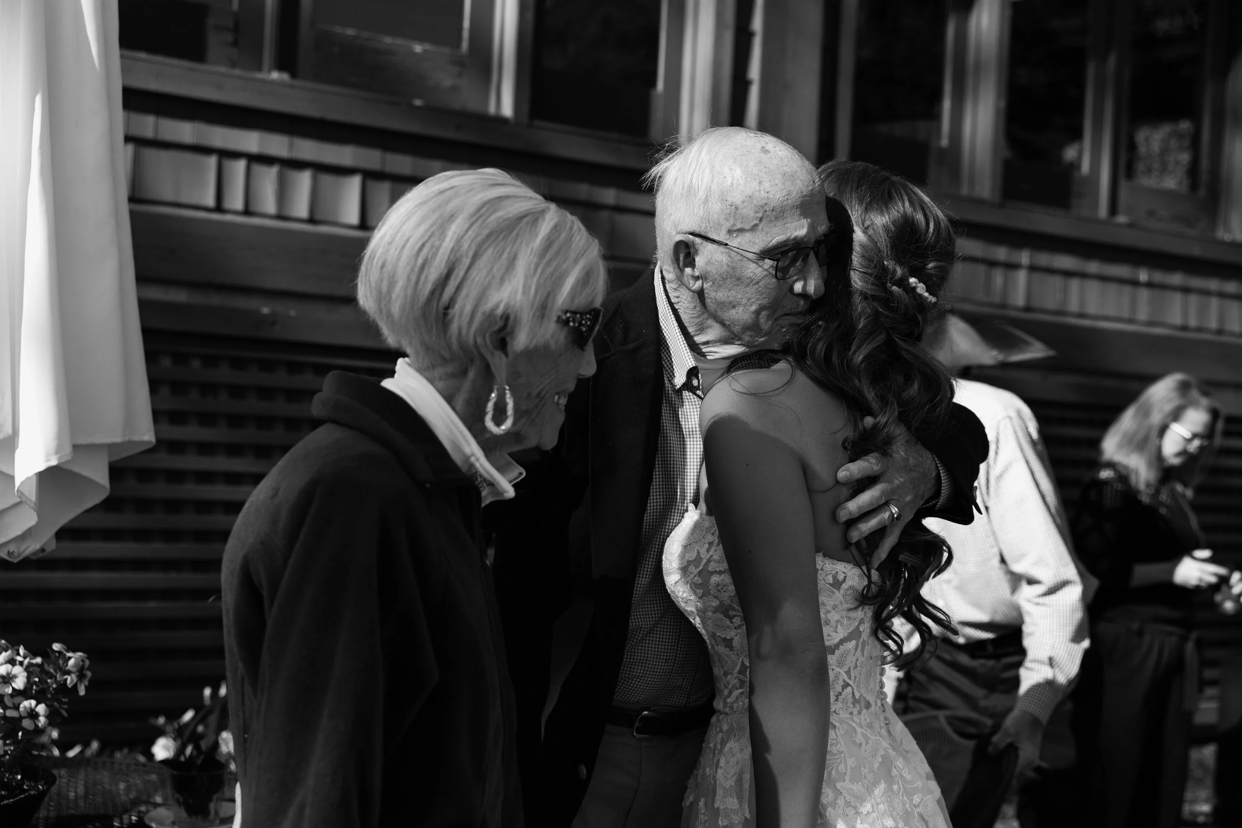 An elderly woman, a man, and a young woman are sitting closely together, sharing a tender moment at a social gathering. The elderly woman has short, light-colored hair and wears glasses, the man has white hair and glasses, and the young woman has lon