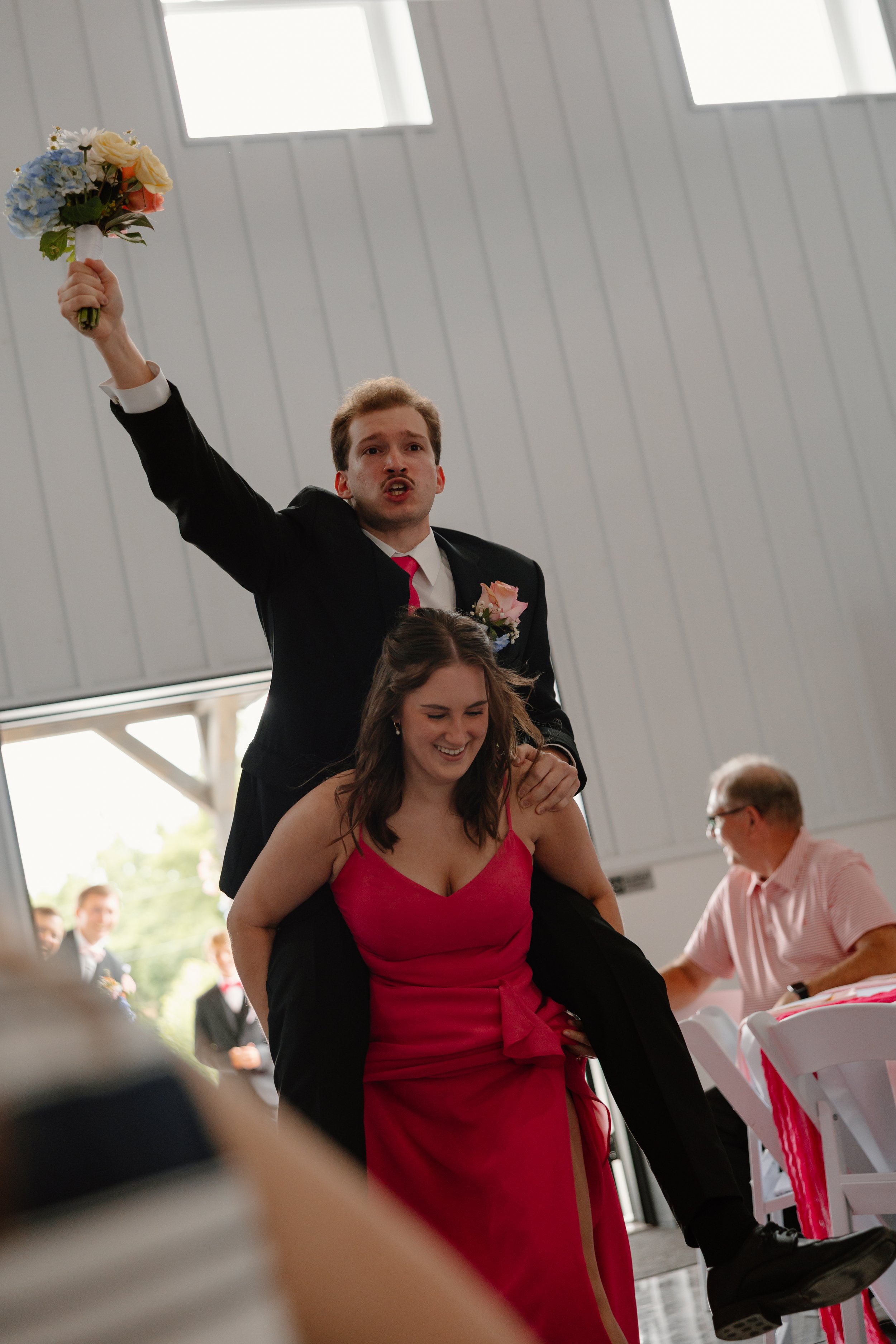 A young man in formal attire, holding a bouquet of flowers, is standing behind a smiling young woman in a red dress, sitting at a decorated table during a celebration or ceremony.
