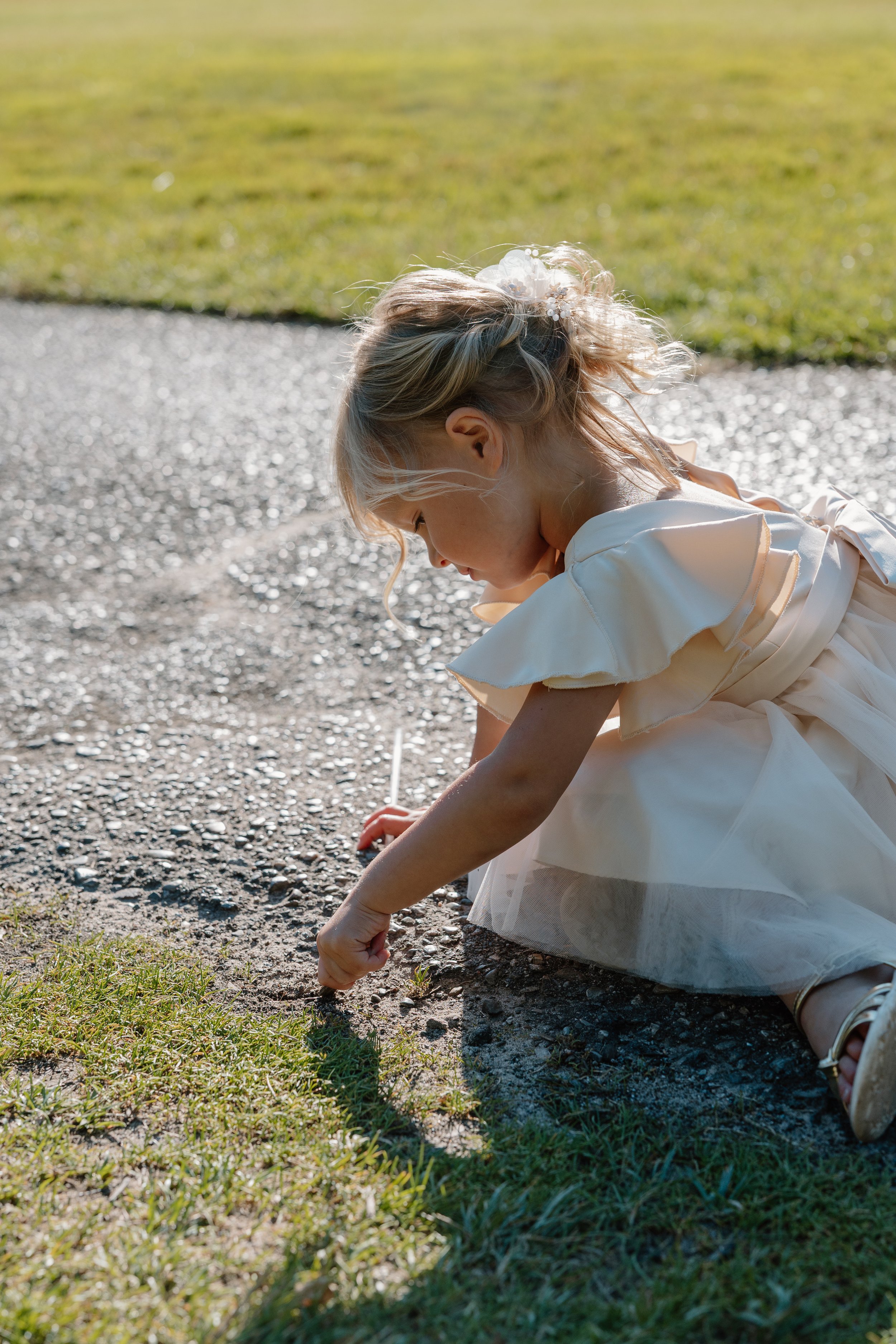 Young girl in a white dress crouches on a gravel path, planting a small seedling or flower in the grass beside the path, with sunlight illuminating her and the surrounding area.