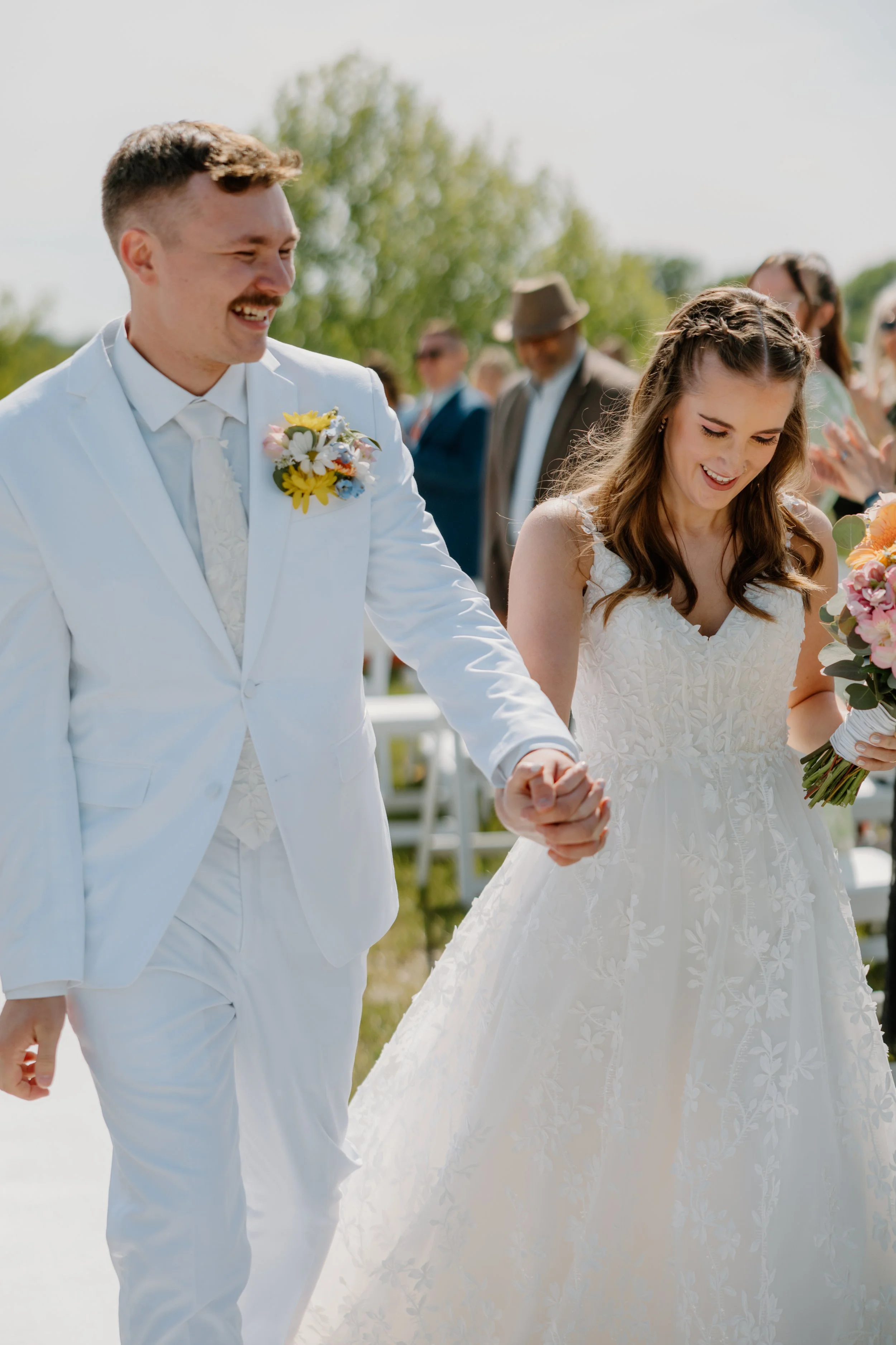 A smiling couple, a man in a white suit with a boutonnière and a woman in a white wedding dress, holding hands during their outdoor wedding.