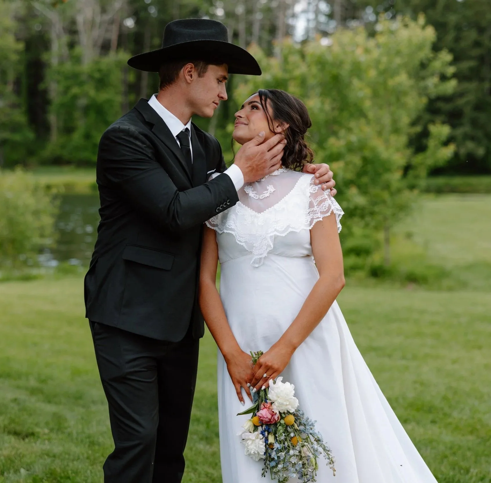 A man and woman dressed in wedding attire standing outdoors by a body of water, with the man gently holding the woman's face while she holds a bouquet of flowers, gazing into each other's eyes.