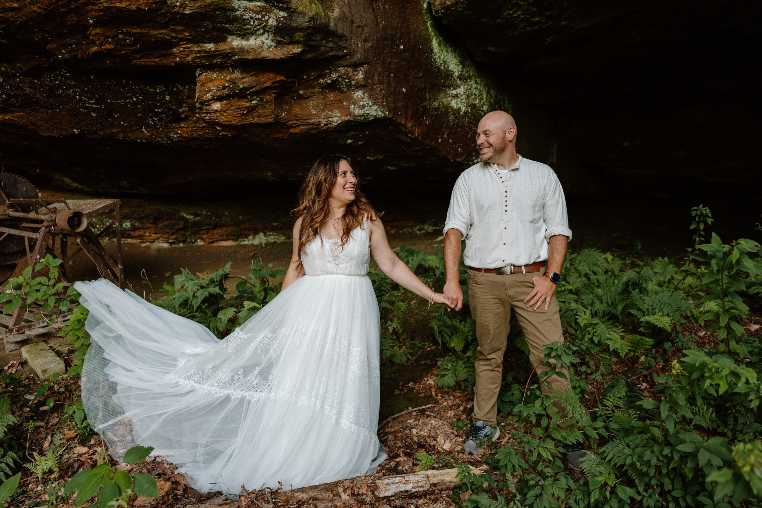 A couple holding hands, smiling, and looking at each other, standing inside a cave surrounded by greenery, with the woman in a white wedding dress and the man in casual clothing.