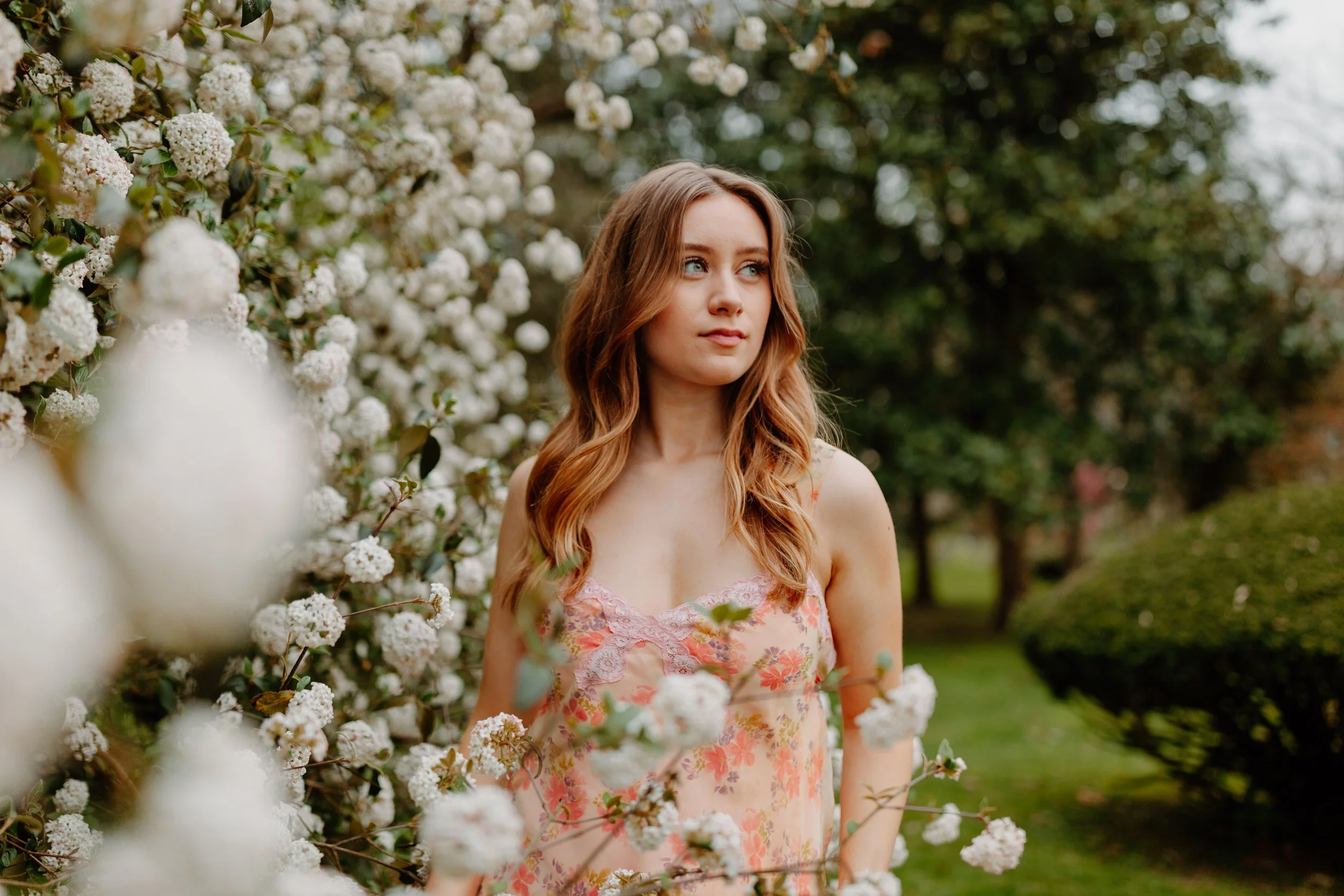 A young woman with long, wavy, reddish-brown hair and blue eyes standing outdoors near a large bush of white flowers, wearing a pink floral dress with lace details.