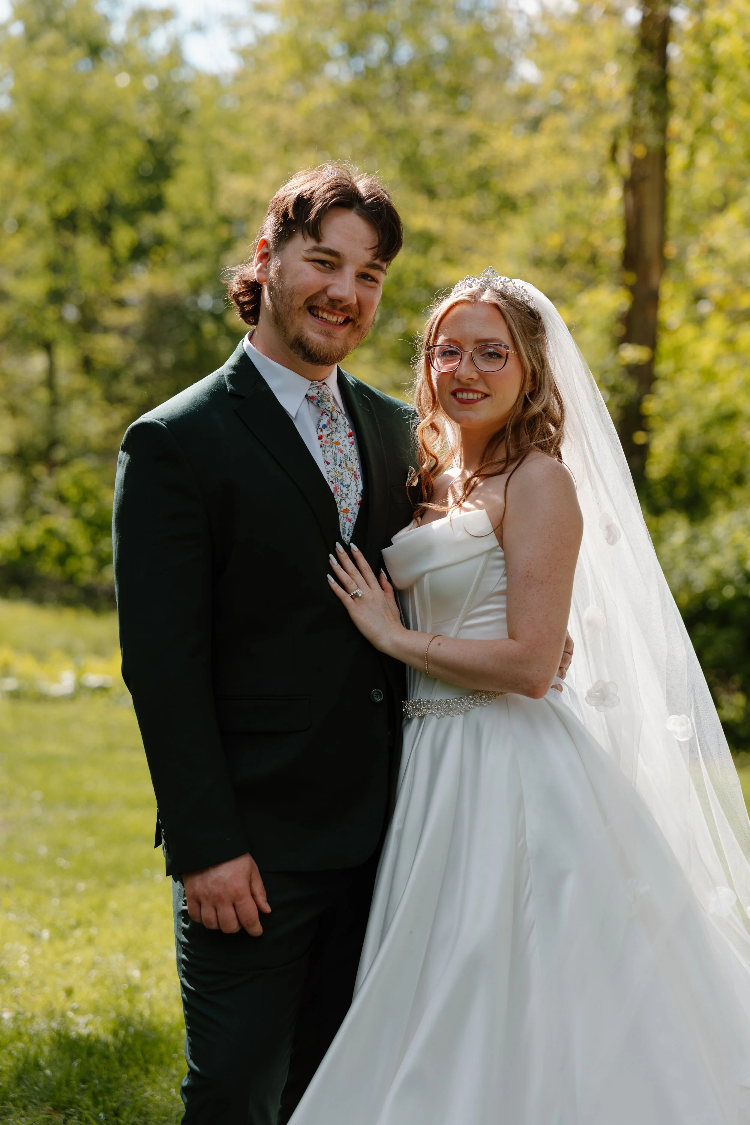 A newlywed couple smiling outdoors in a park with green trees, the groom in a black suit and tie, and the bride in a white wedding gown with a veil and tiara.