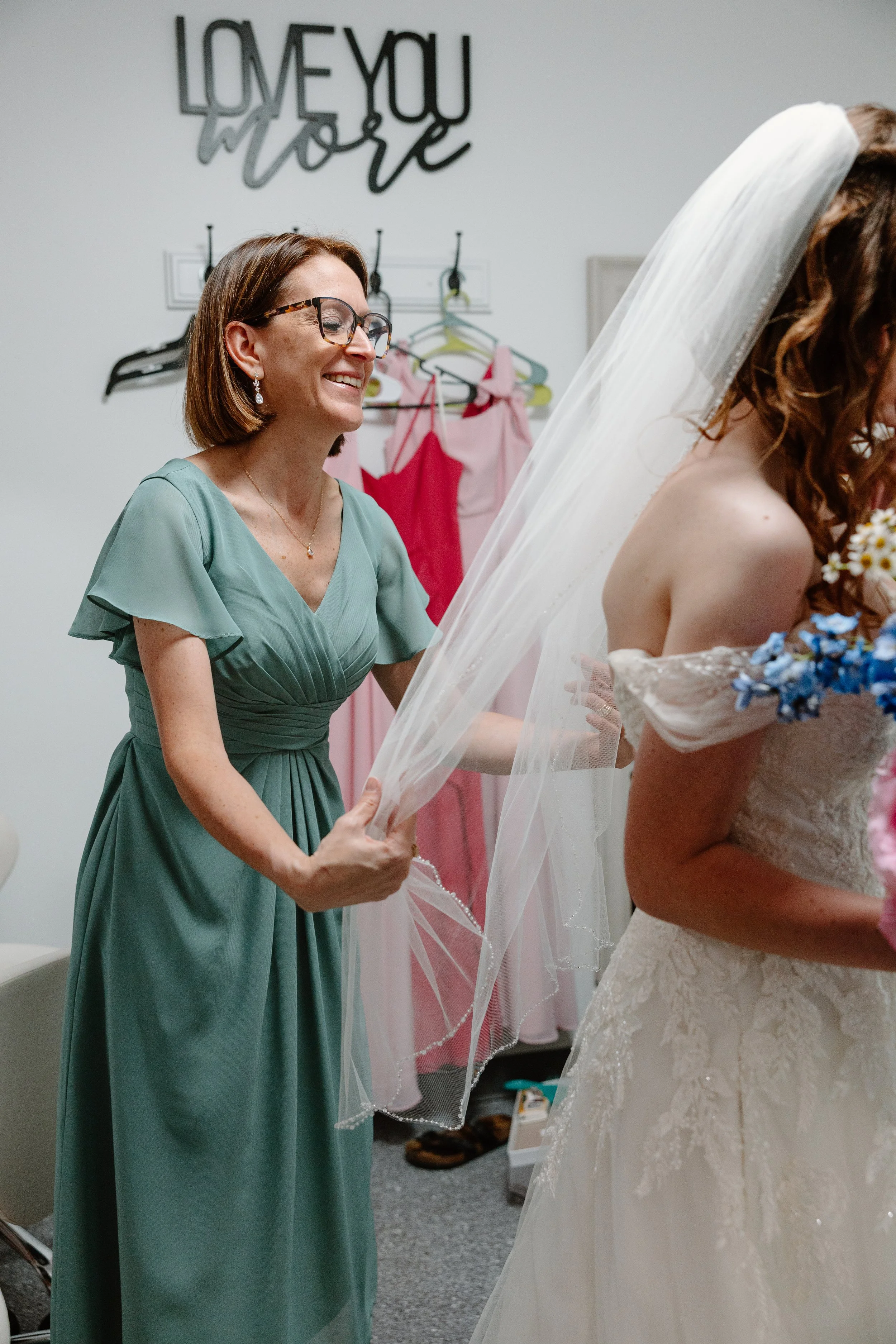 A woman in a green dress holding a veil in a dressing room, smiling at a bride in a wedding gown holding a bouquet of flowers. The background has a sign that reads 'Love You More' and pink dresses hanging on hooks.
