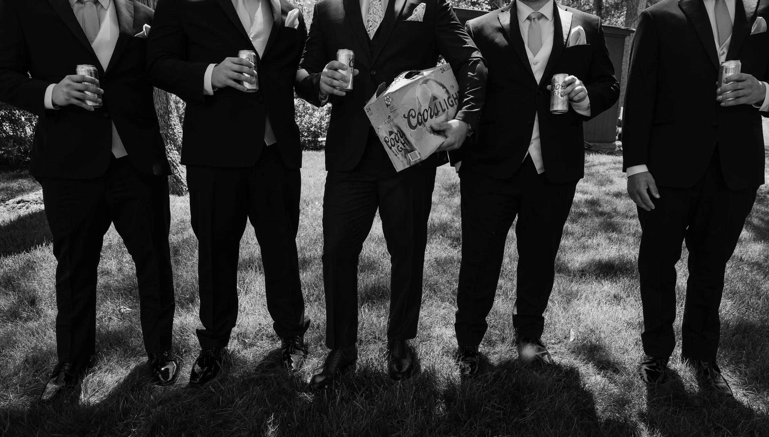 Six men dressed in suits standing outdoors on grass, holding cans of Coors Light beer. One man in the center is holding a six-pack of beer, while the others are holding individual cans.