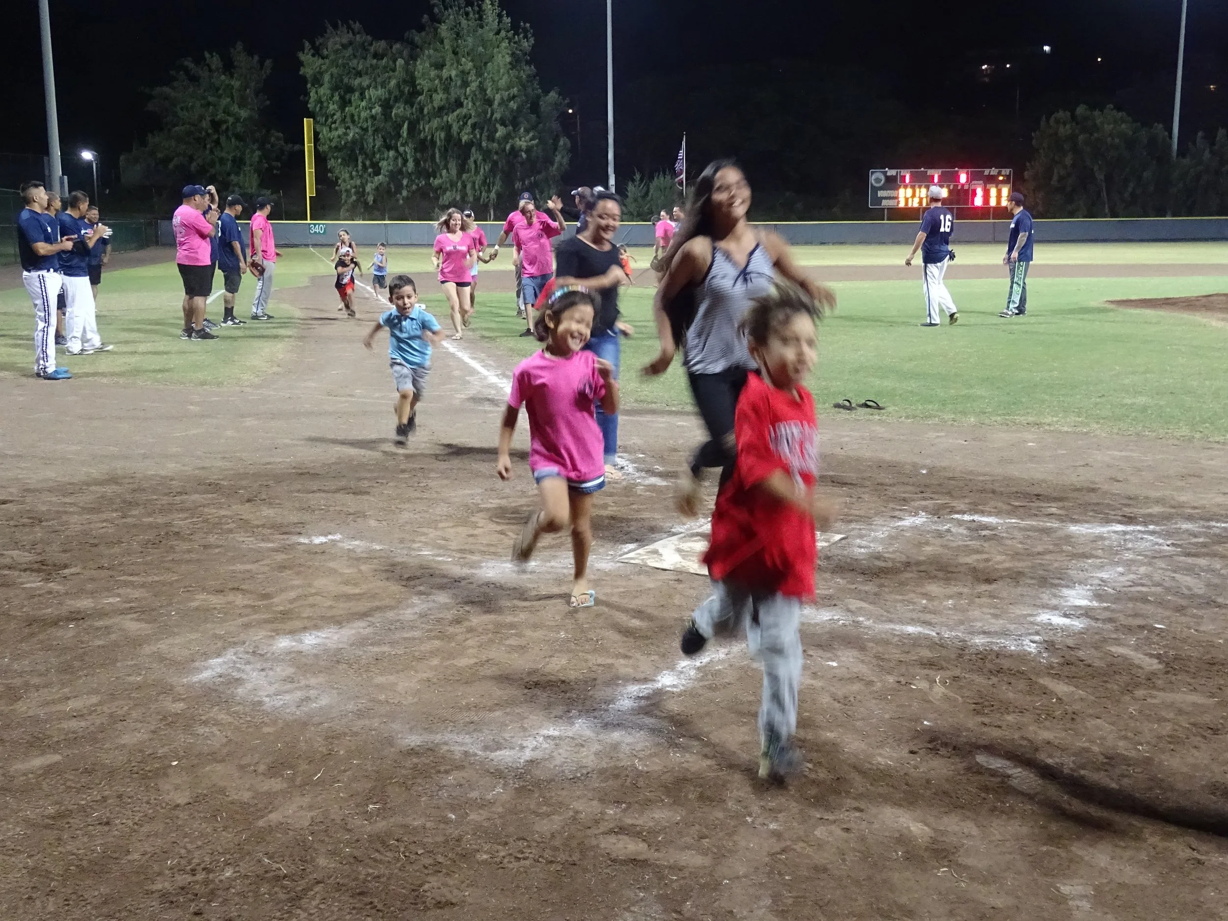 People, including children and adults, running on a baseball field at night, with some of them smiling and having fun, illuminated by stadium lights.