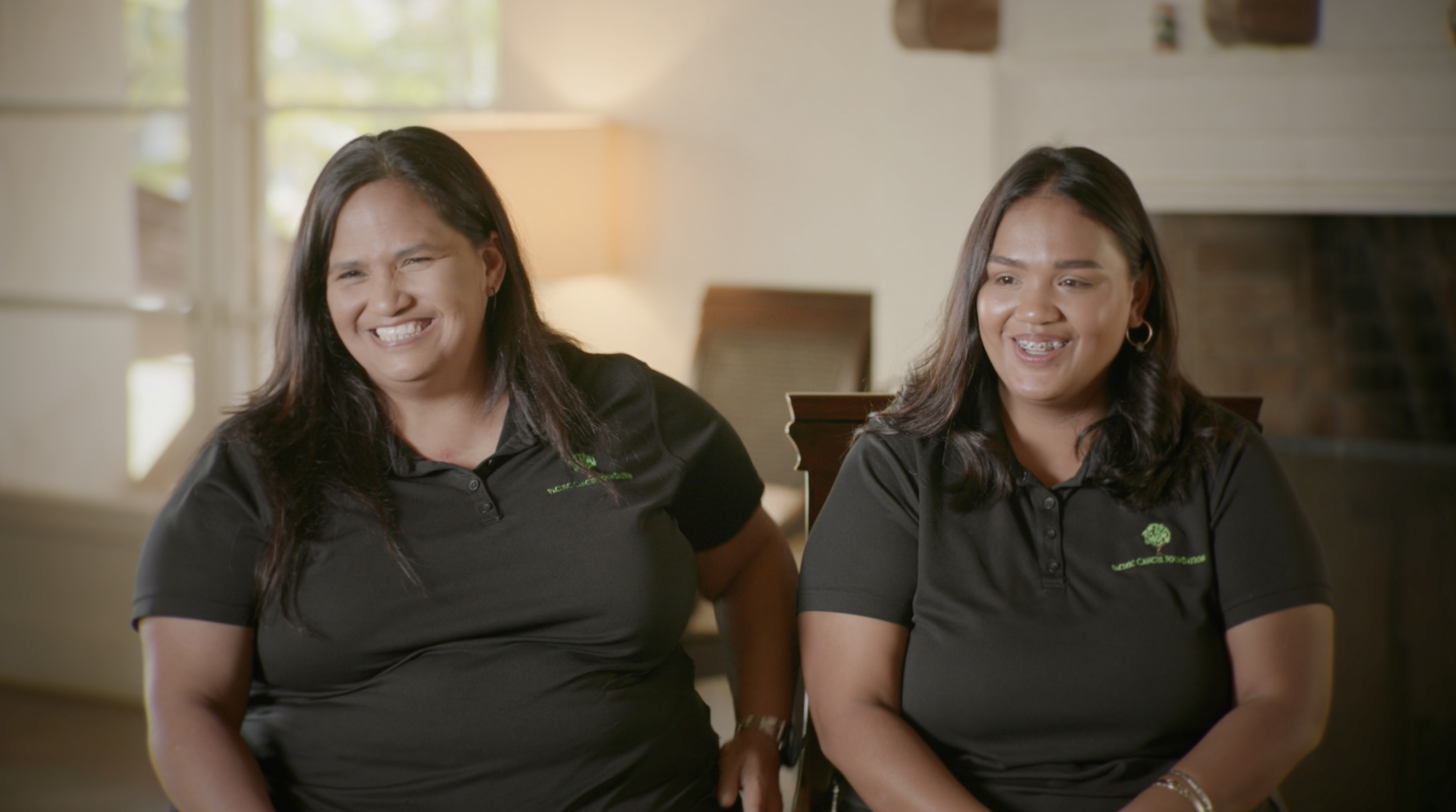 Two women sitting side by side, smiling, wearing black polo shirts with green logos that read 'Pacific Cancer Foundation' on the left chest, in a warmly lit room with windows and a lamp in the background.