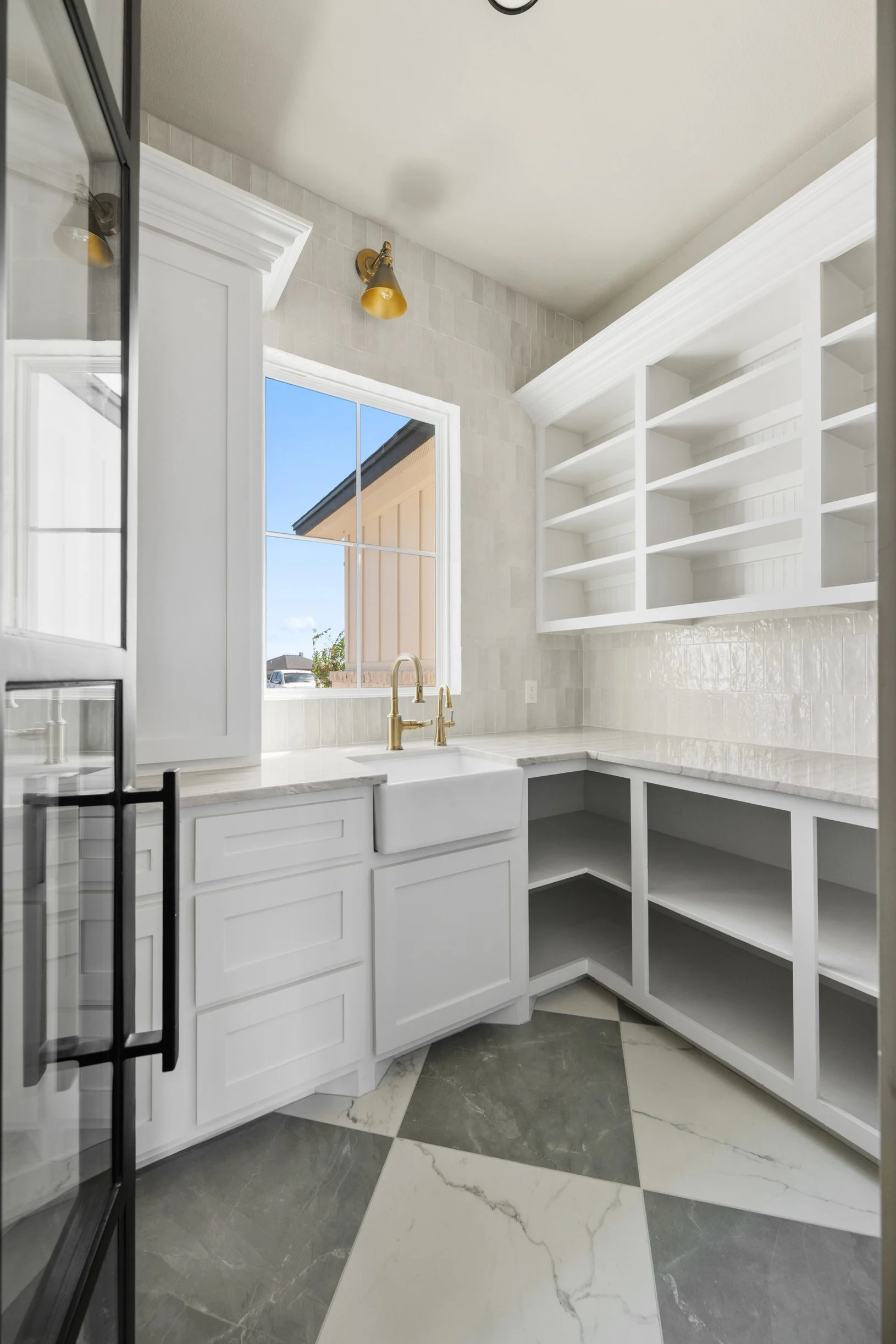 Modern kitchen with white cabinets, open shelving, marble countertops, and a large window letting in natural light. There is a gold-colored faucet and a tiled backsplash. The floor has a checkered pattern with black and white marble tiles.