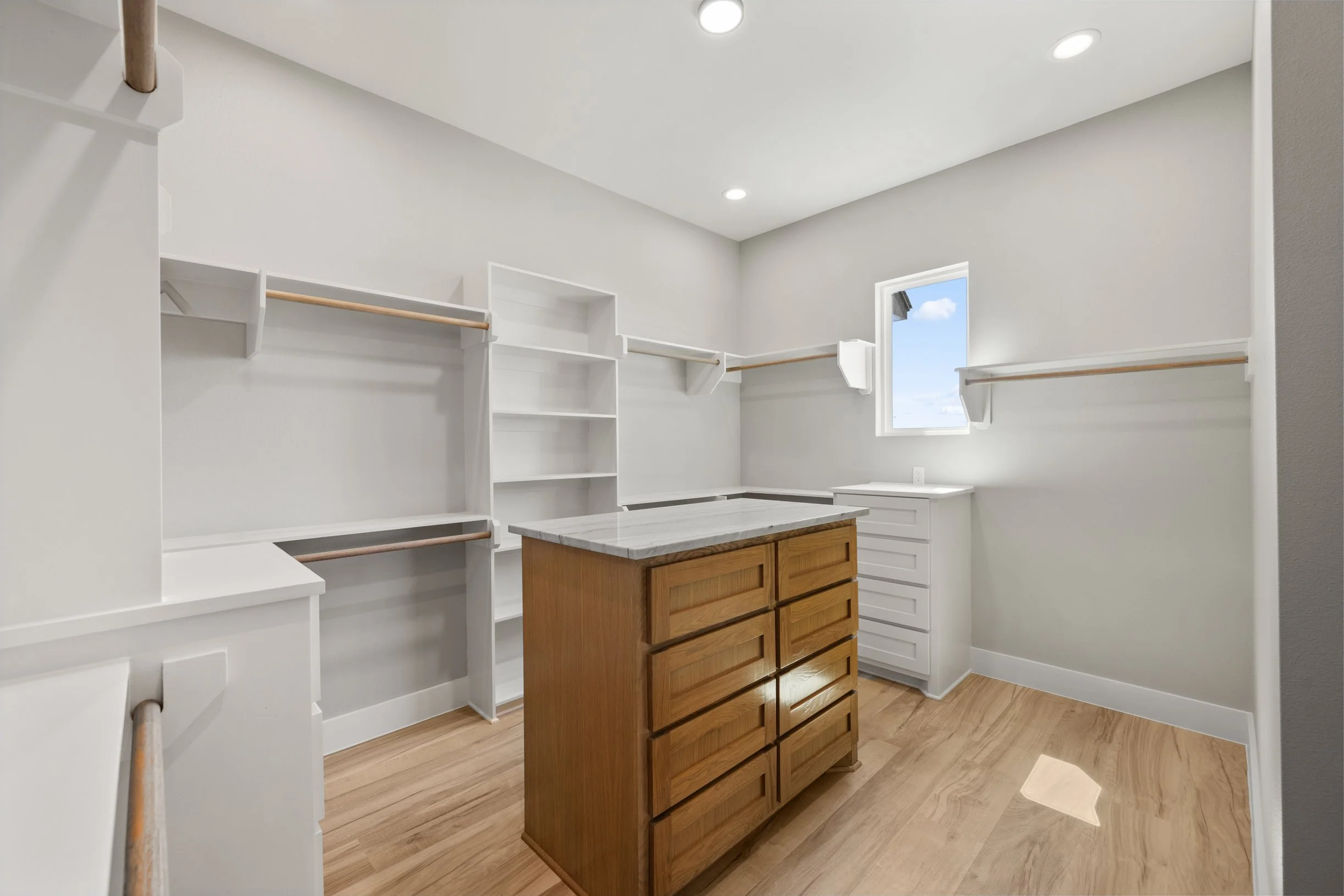 Empty walk-in closet with white shelves, rods for clothing, a small white cabinet, a wooden island with drawers, and a window showing the sky outside.