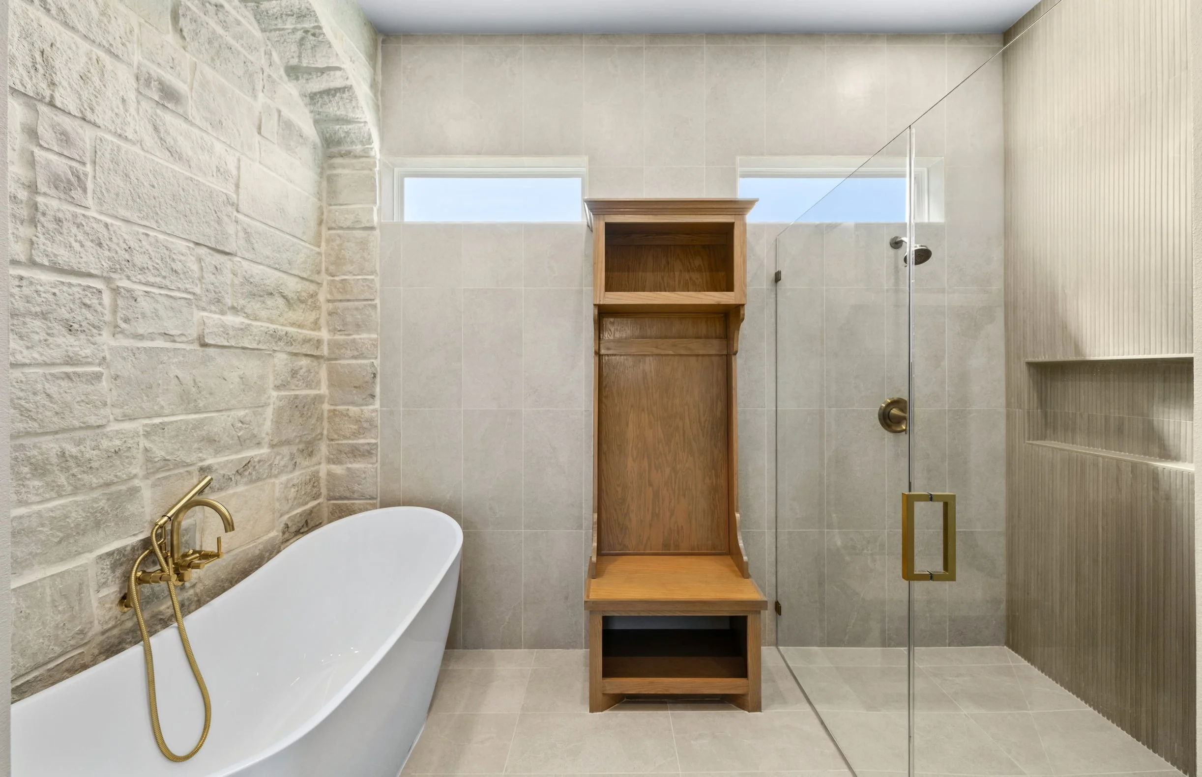 A modern bathroom with a white bathtub, brass fixtures, a stone accent wall, beige tiles, a wooden storage unit, and a glass shower enclosure with a gold handle.