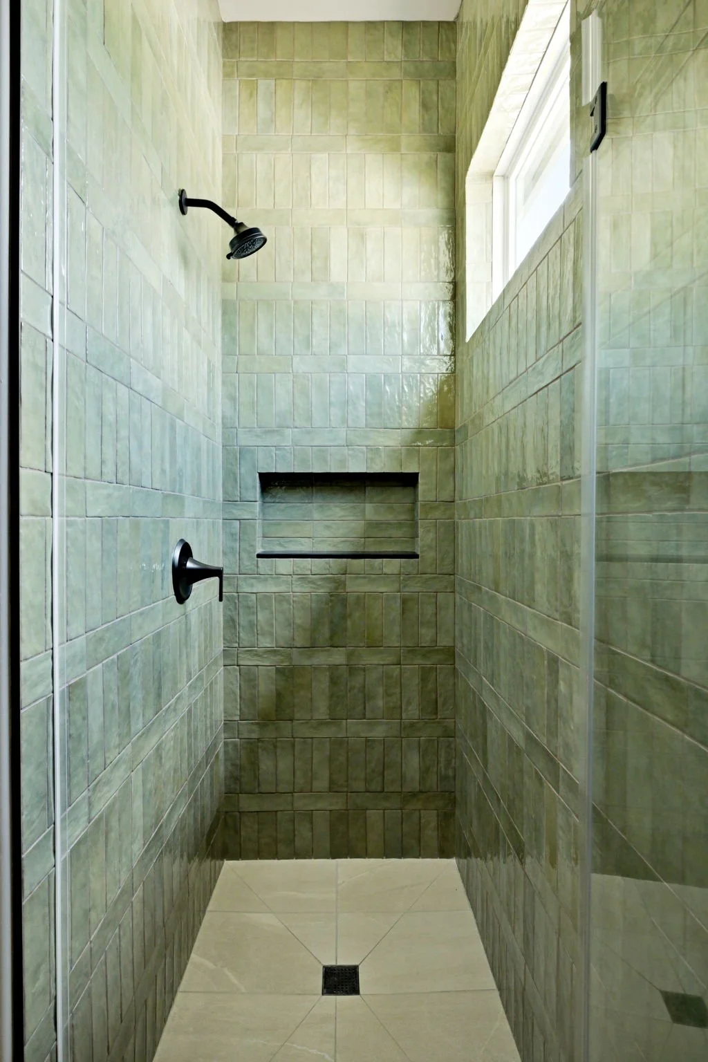 A walk-in shower with green tile walls, a black showerhead, a black handle, a built-in niche for toiletries, a window for natural light, and a beige tiled floor with a floor drain.