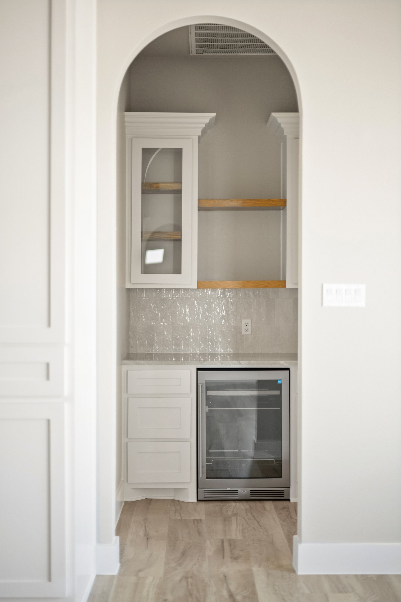 Interior view of a built-in kitchen area with white cabinetry, wooden countertops, a glass-fronted refrigerator, and a small bar fridge below, framed by an arched doorway with marble flooring.